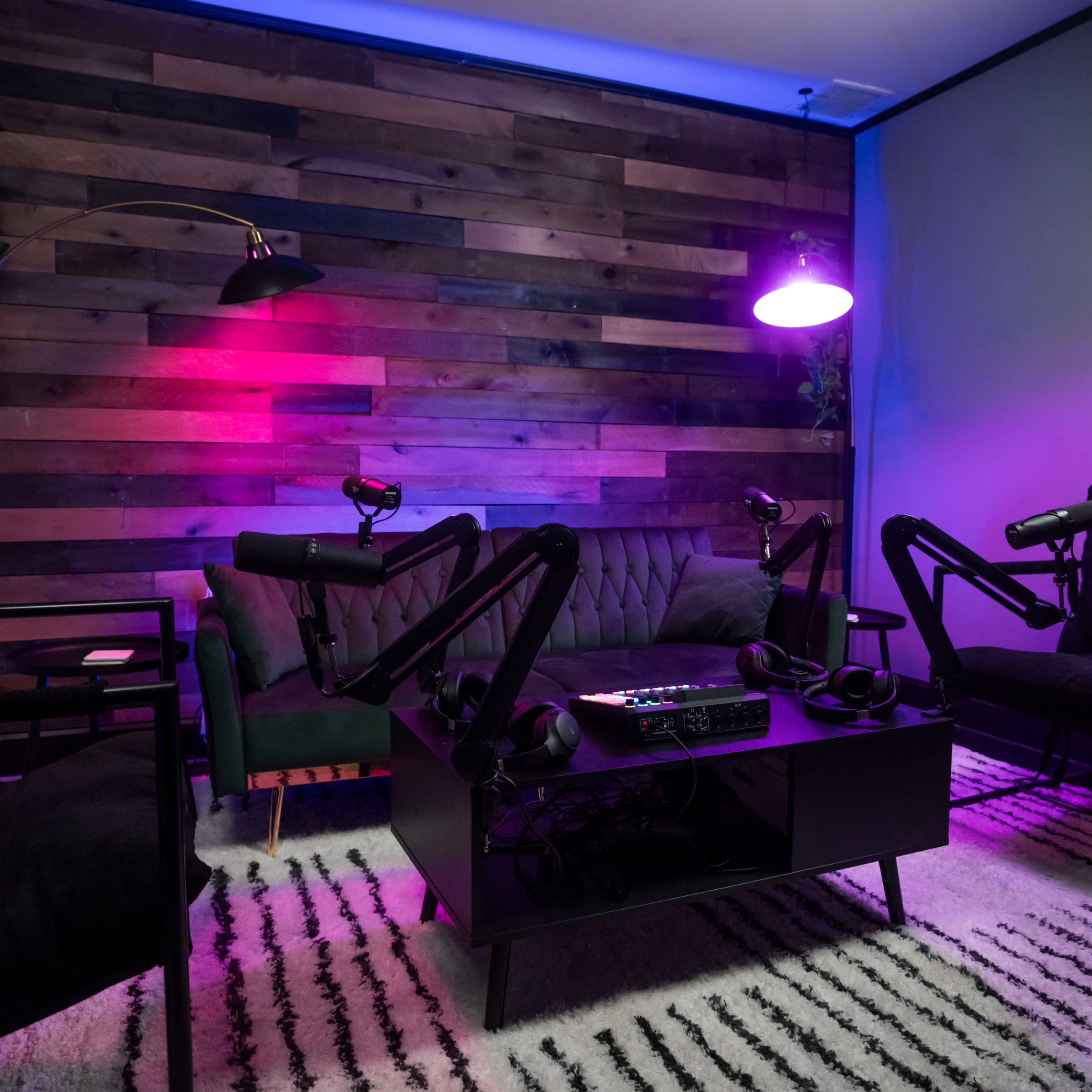 A modern podcasting studio features a wooden accent wall, four chairs, and audio equipment, illuminated by purple and pink lighting.