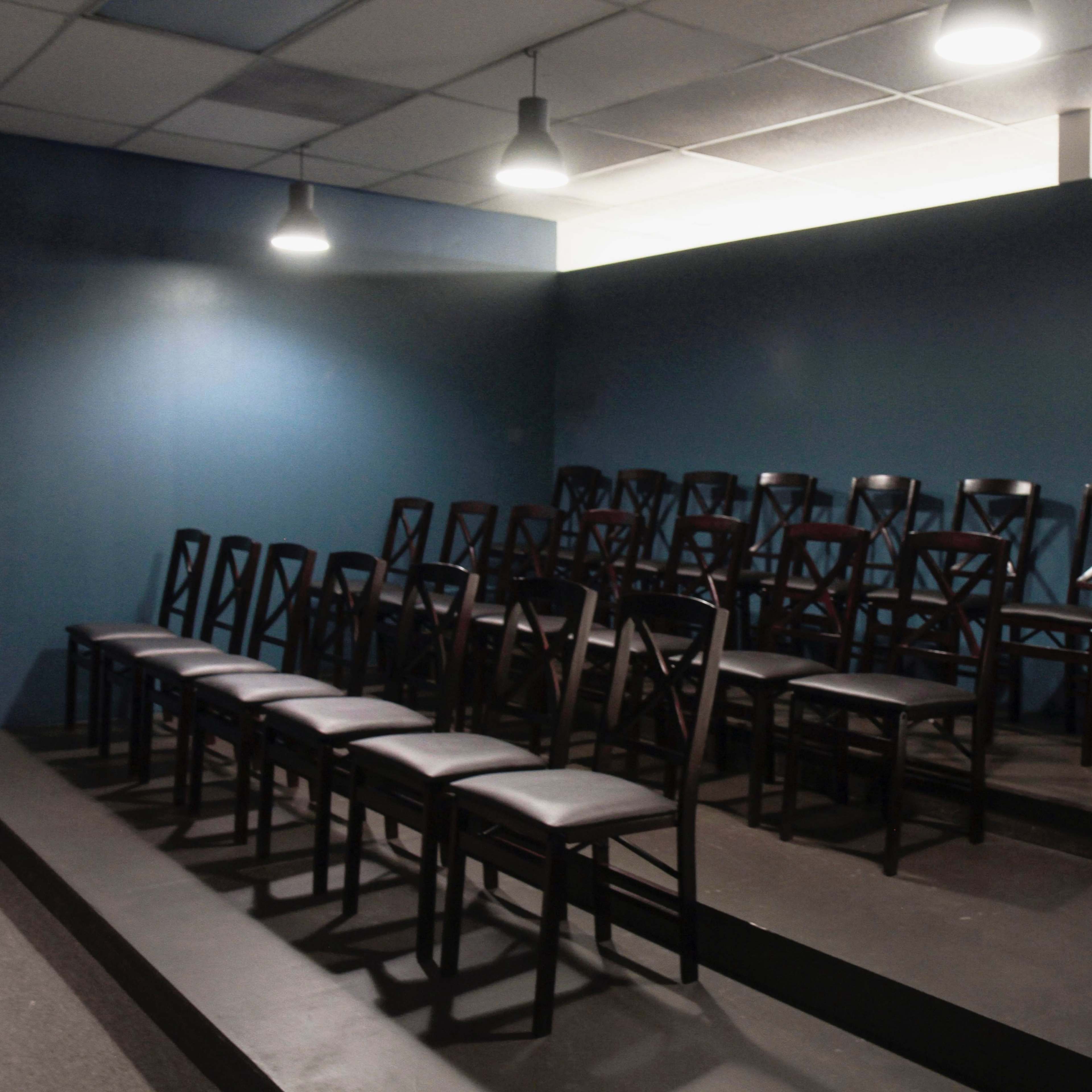 A set of black wooden chairs is arranged in rows on a raised platform against a blue wall under overhead lights.