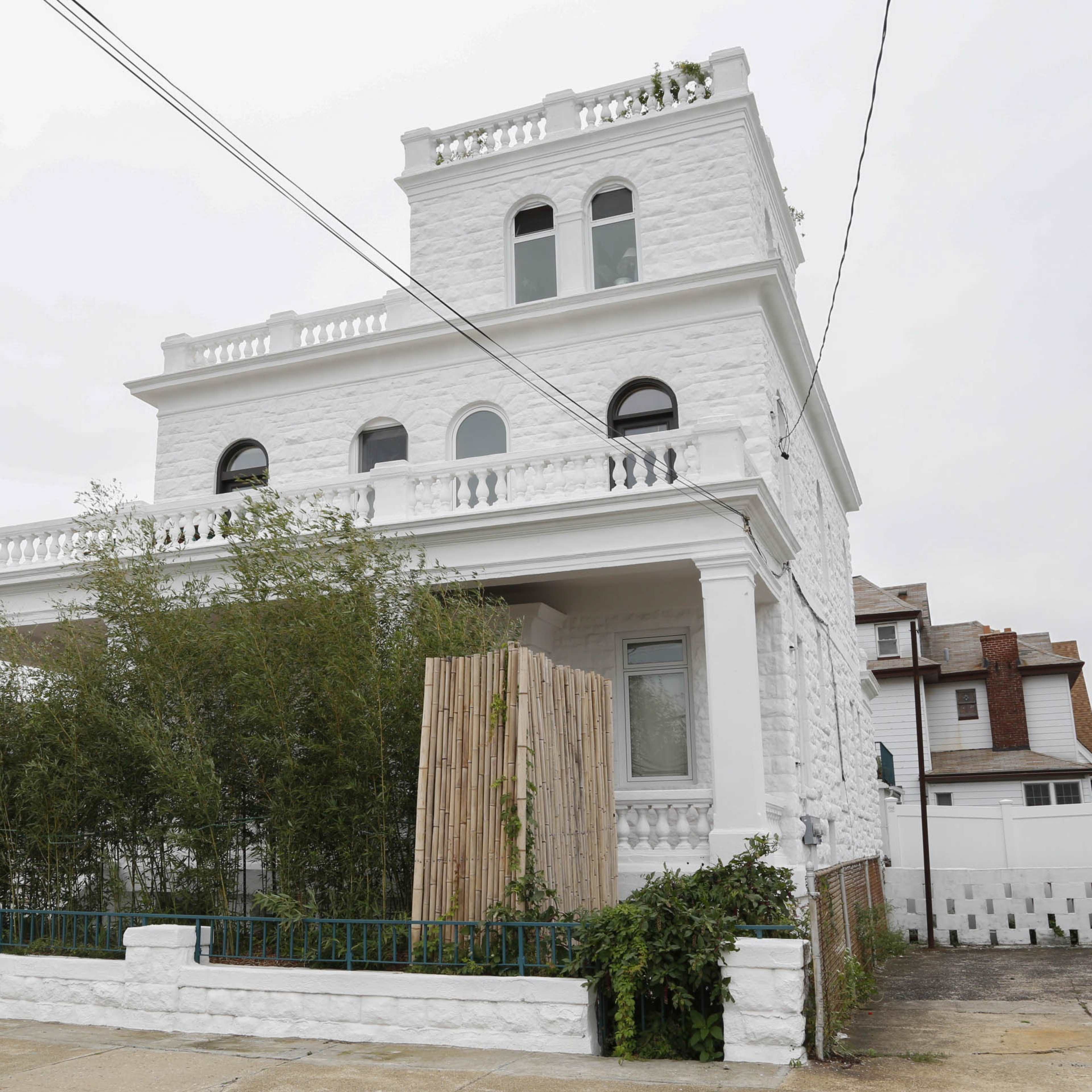 A large, white, three-story house features a balcony and a bamboo fence in the front yard.