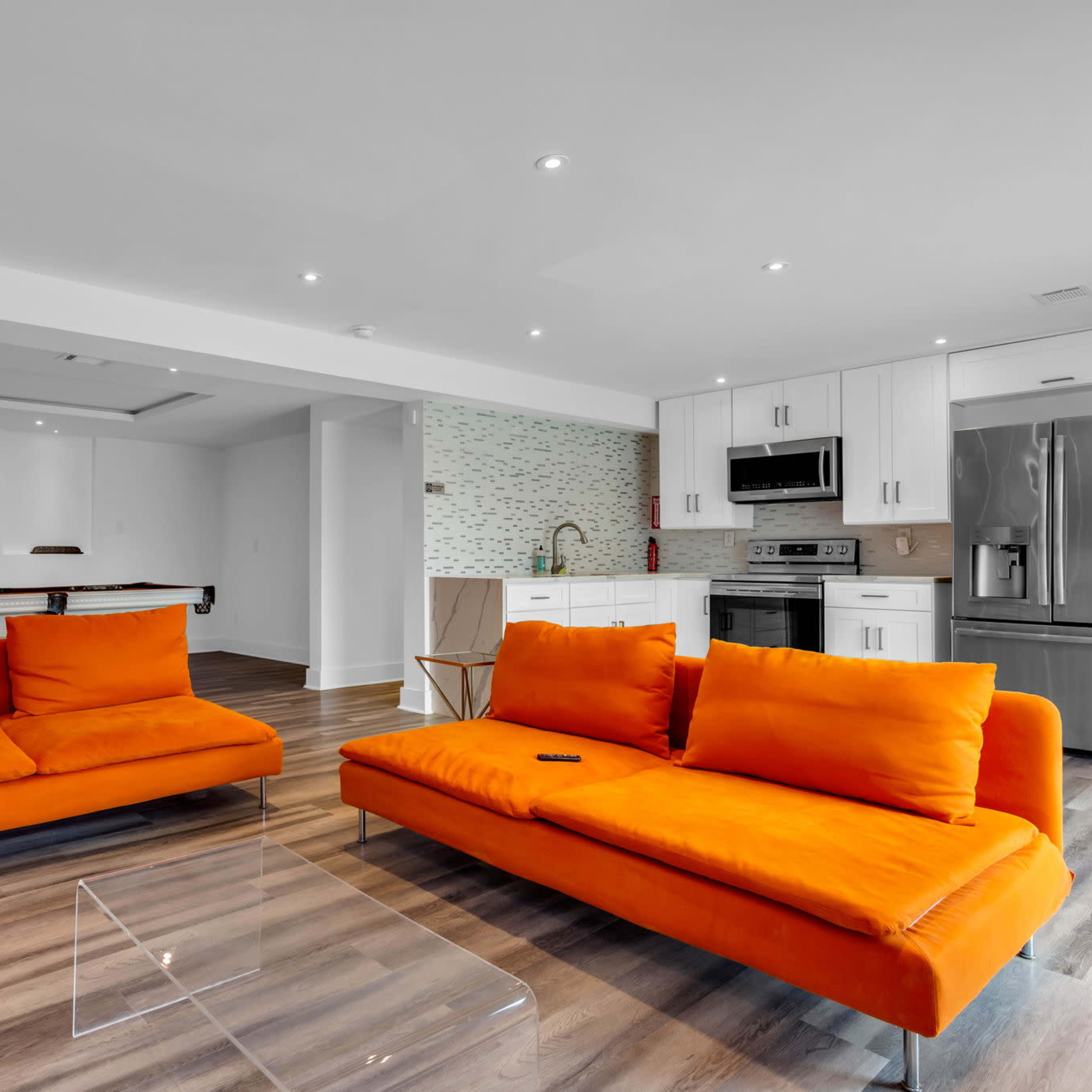 A modern living space features two orange sofas facing a kitchen with stainless steel appliances and a pool table in the background.