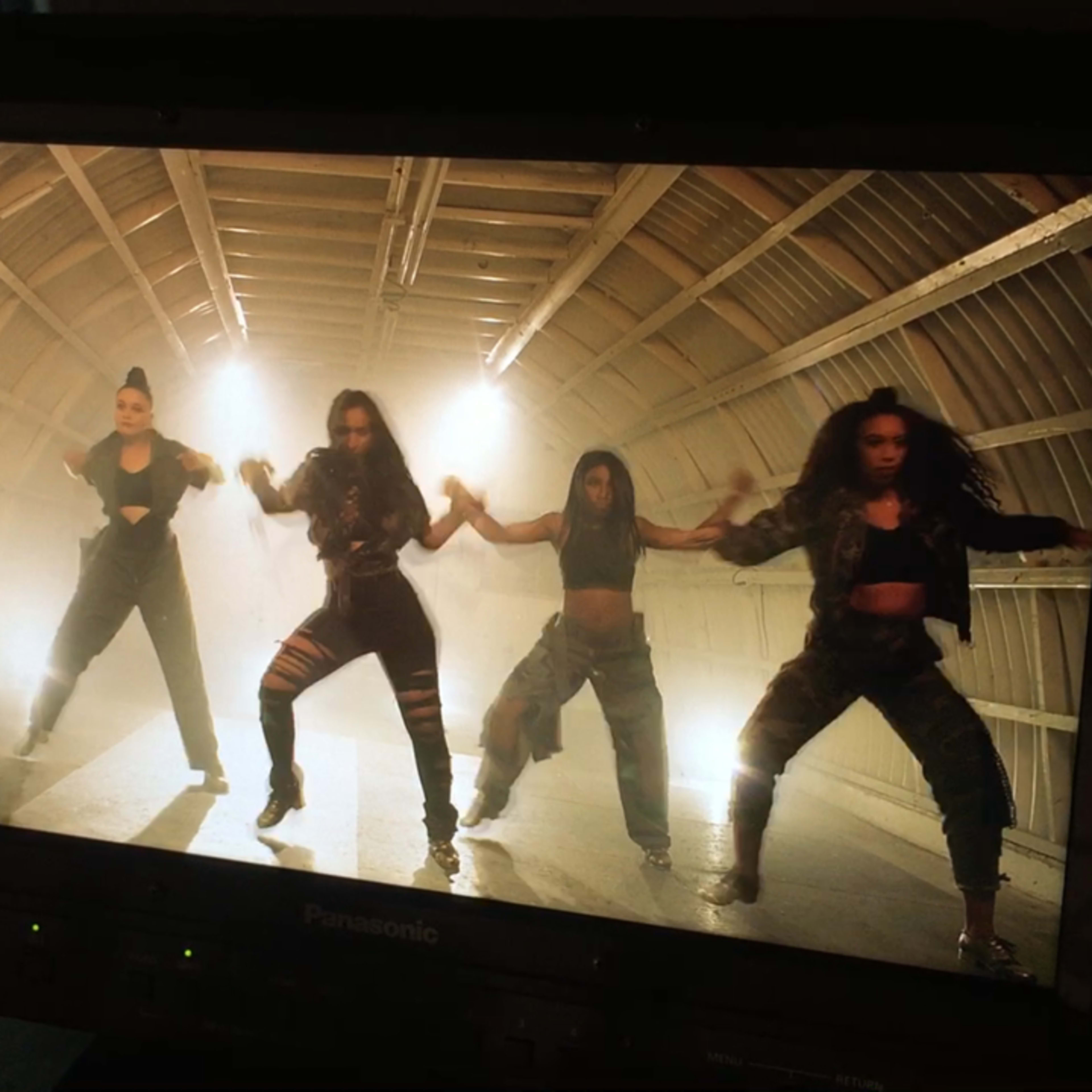 A group of five dancers performs synchronized movements in a dimly lit studio with a wooden ceiling.
