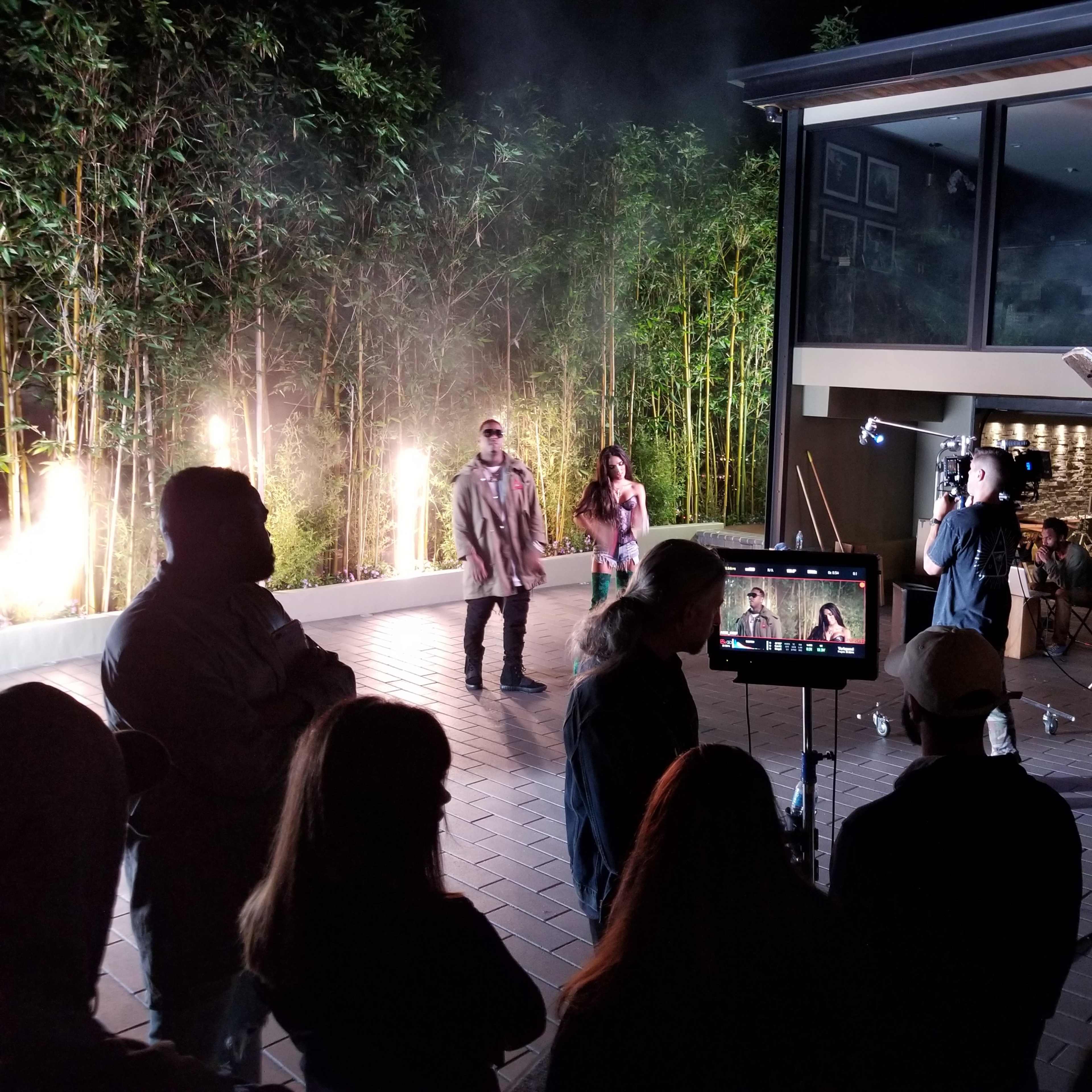 A film crew is filming a scene outdoors near a bamboo backdrop, with lights and equipment set up while several people observe.