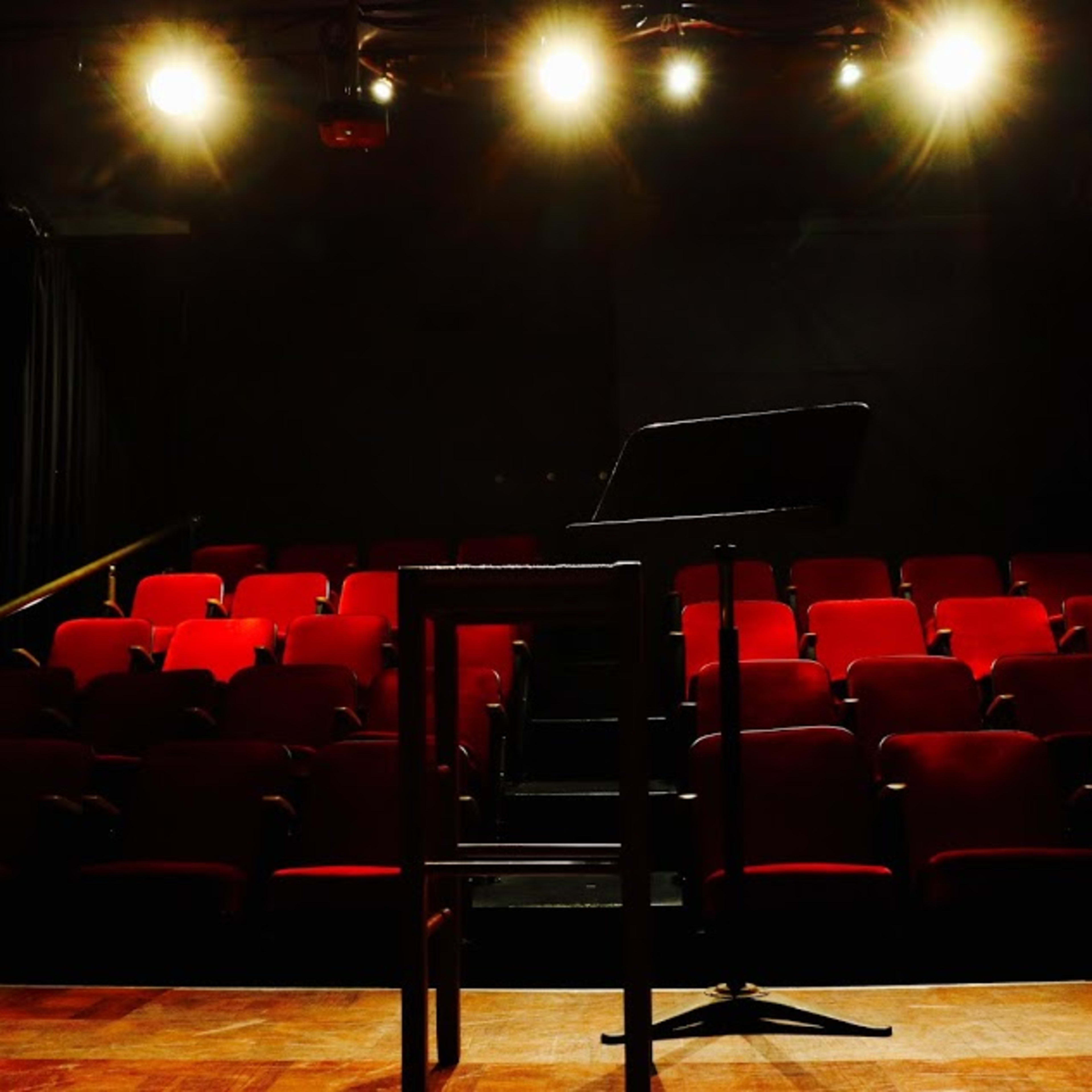 A dimly lit theater stage with a music stand in the foreground and red theater seats arranged in rows behind it.