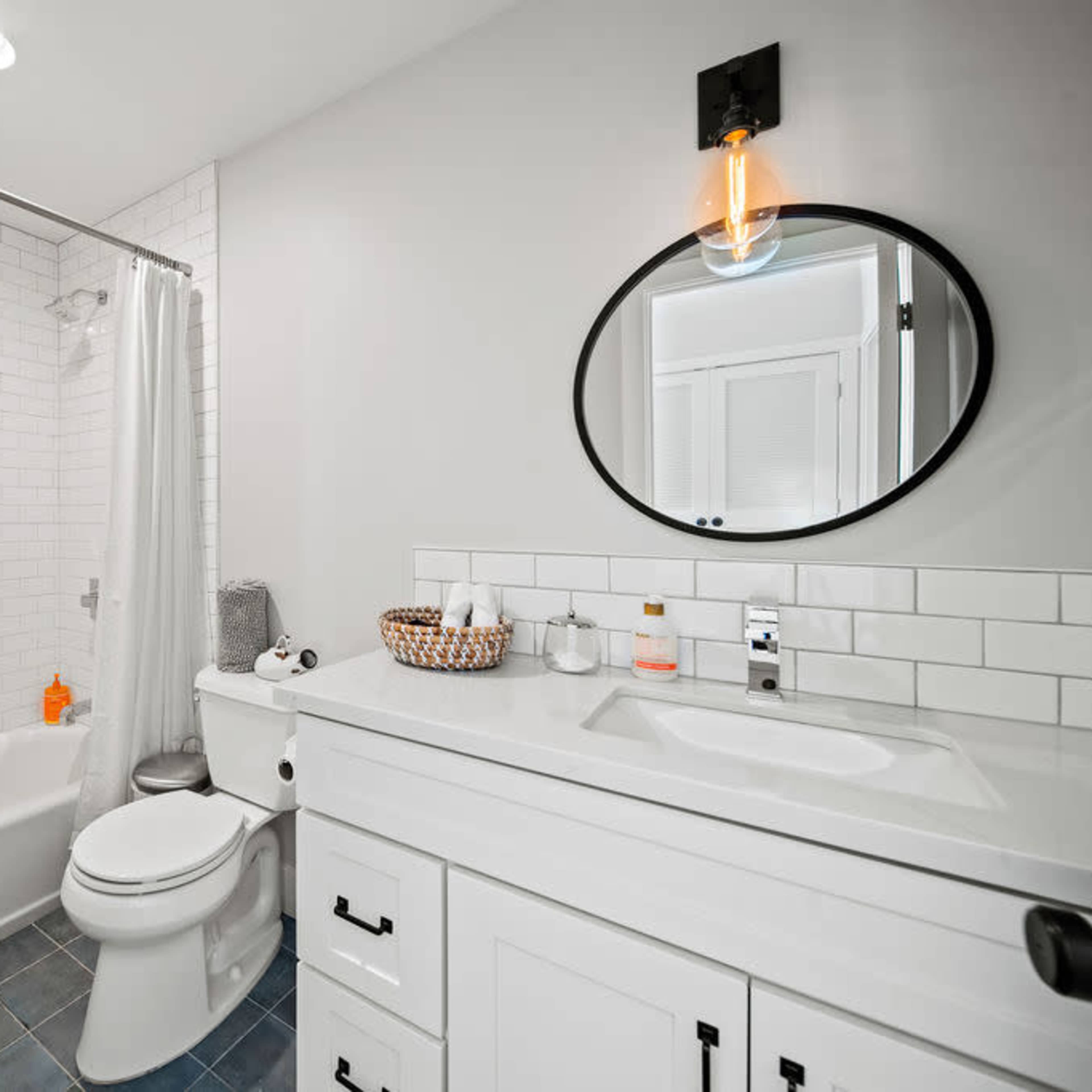 A modern bathroom with a white tiled shower, a light-colored vanity, and a round mirror.