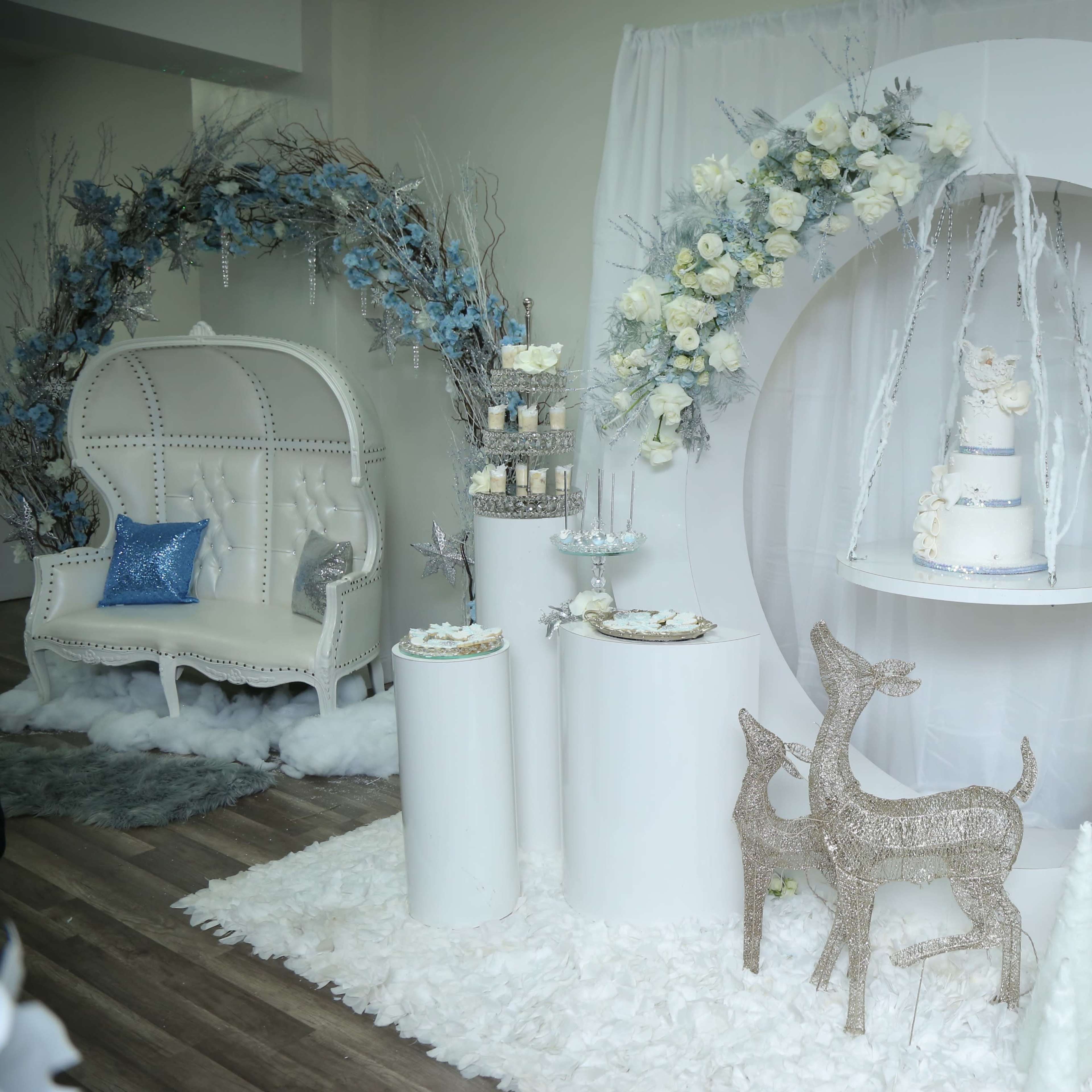 The scene features a decorative event setup with white and blue floral arrangements, a multi-tiered cake on display, and elegant furniture, all arranged against a soft white backdrop.