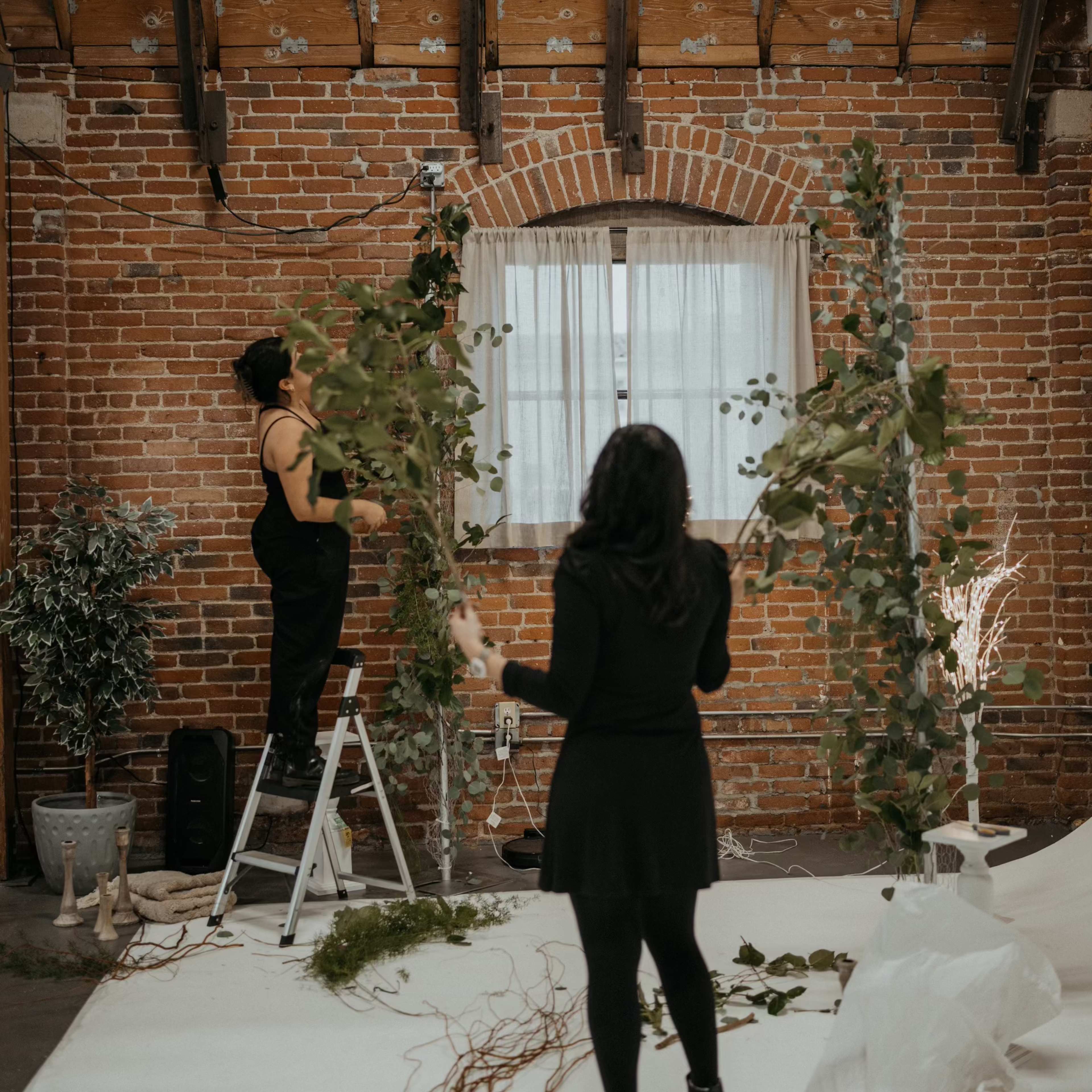 Two people are arranging greenery and decorative elements in a rustic, brick-walled space for an event.