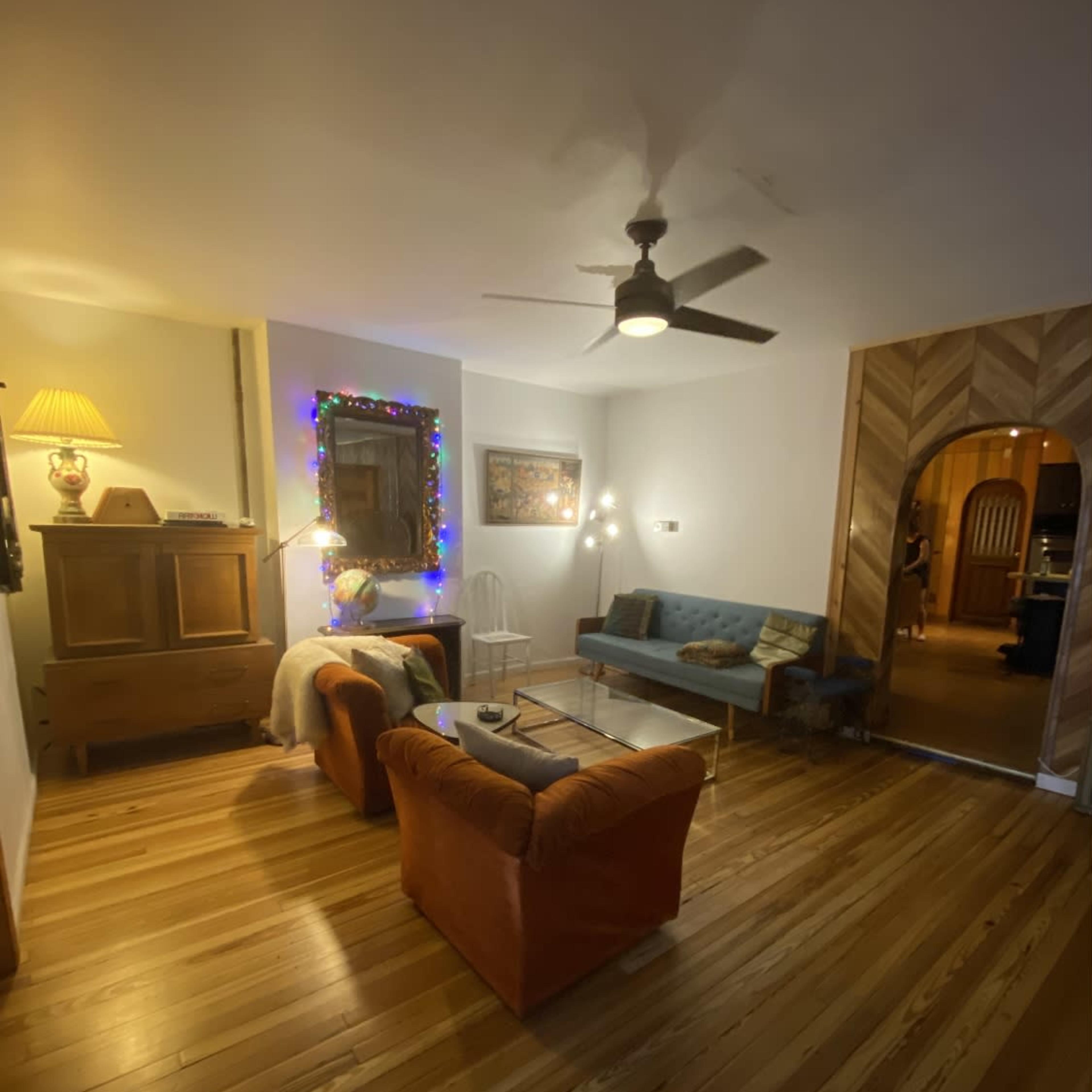 A living room with wooden floors, a blue couch, two orange chairs, and a ceiling fan, illuminated by soft lighting and adorned with decorations.