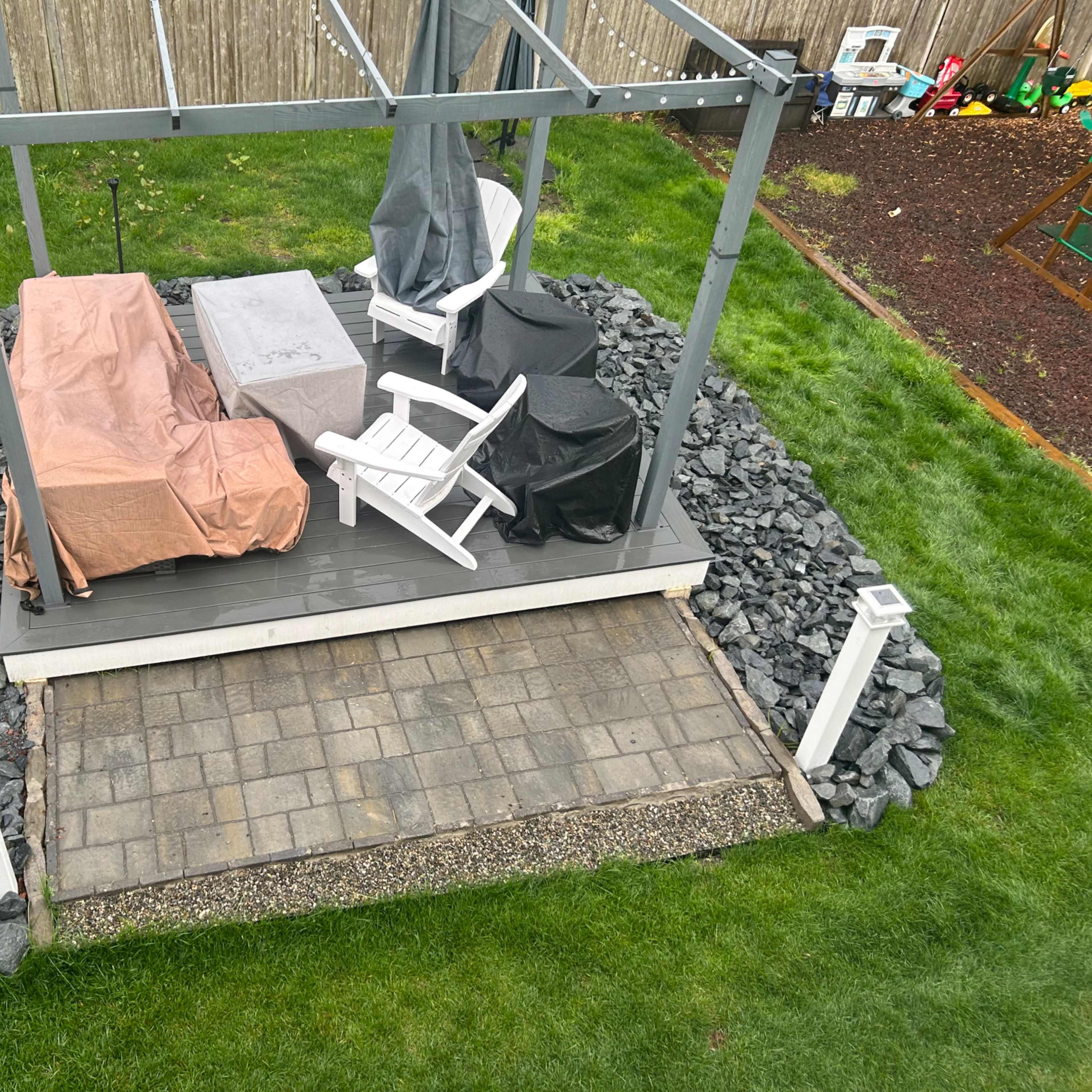 A patio with a covered seating area, featuring white chairs and a table, surrounded by gravel and grass, with a garden bed in the background.