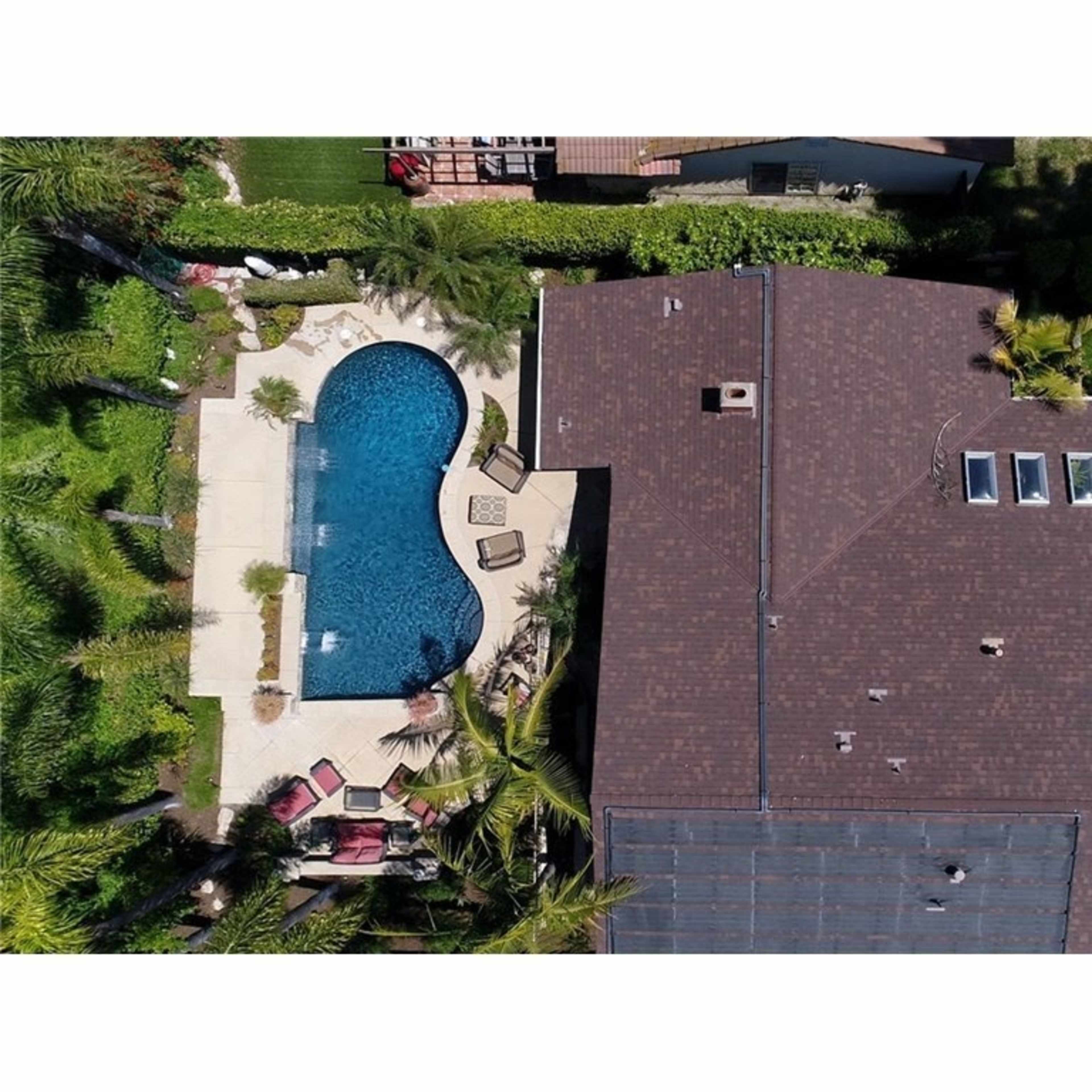 An aerial view shows a house with a brown roof and a backyard featuring a swimming pool surrounded by patio furniture and landscaping.