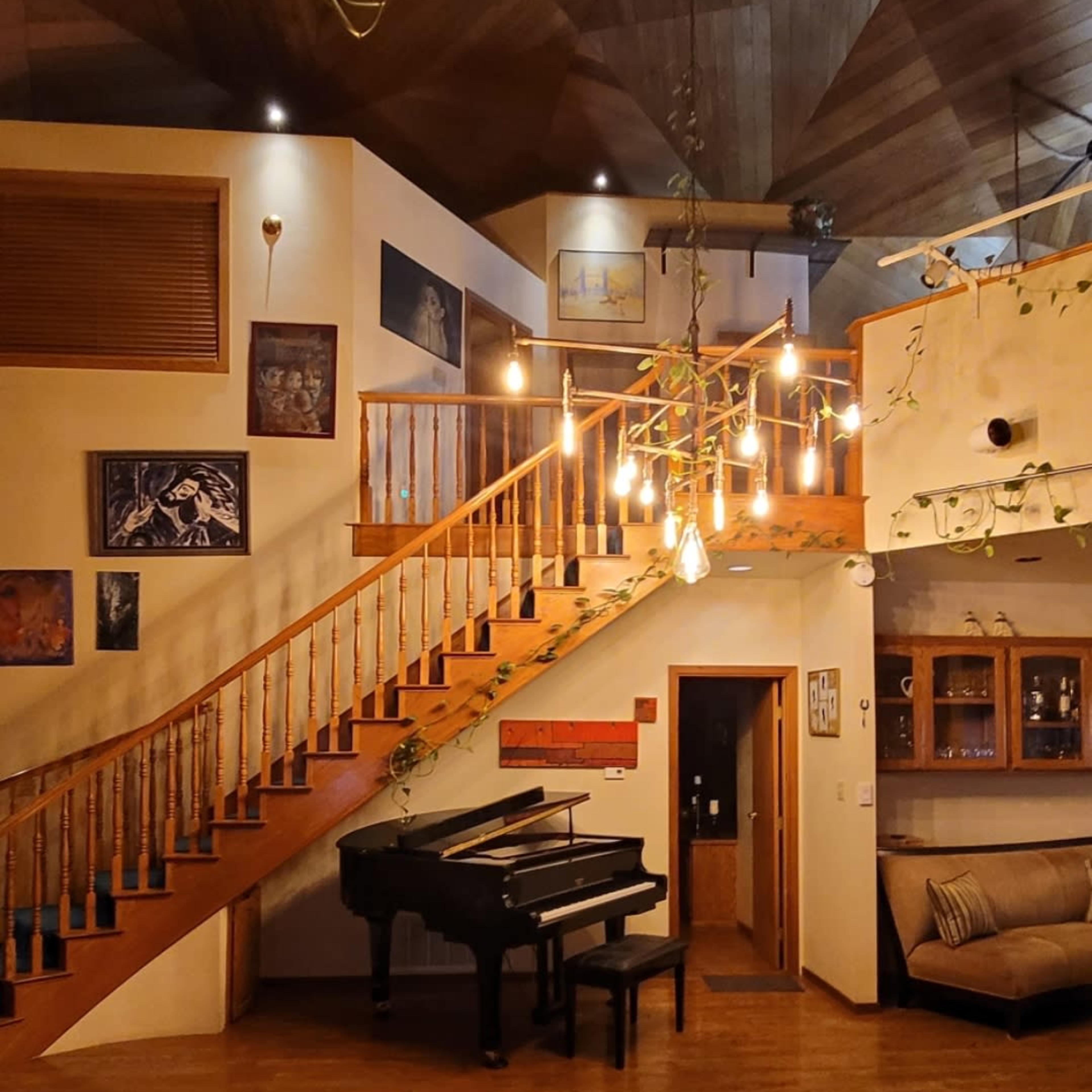 A spacious interior with a grand piano positioned beside a staircase, illuminated by hanging light bulbs and adorned with artwork on the walls.