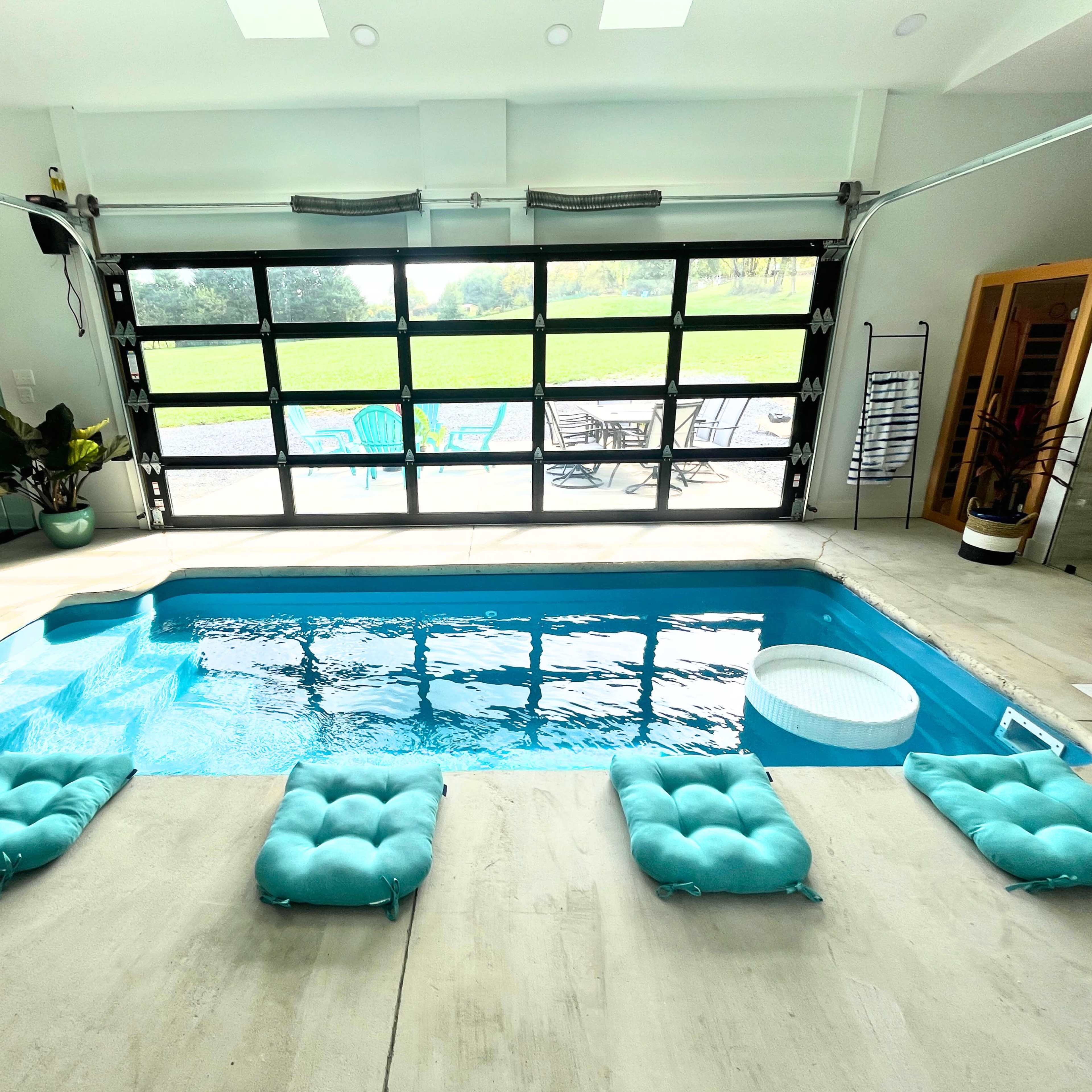 A small indoor pool with turquoise cushions placed around it, surrounded by large glass doors that open to a grassy area.