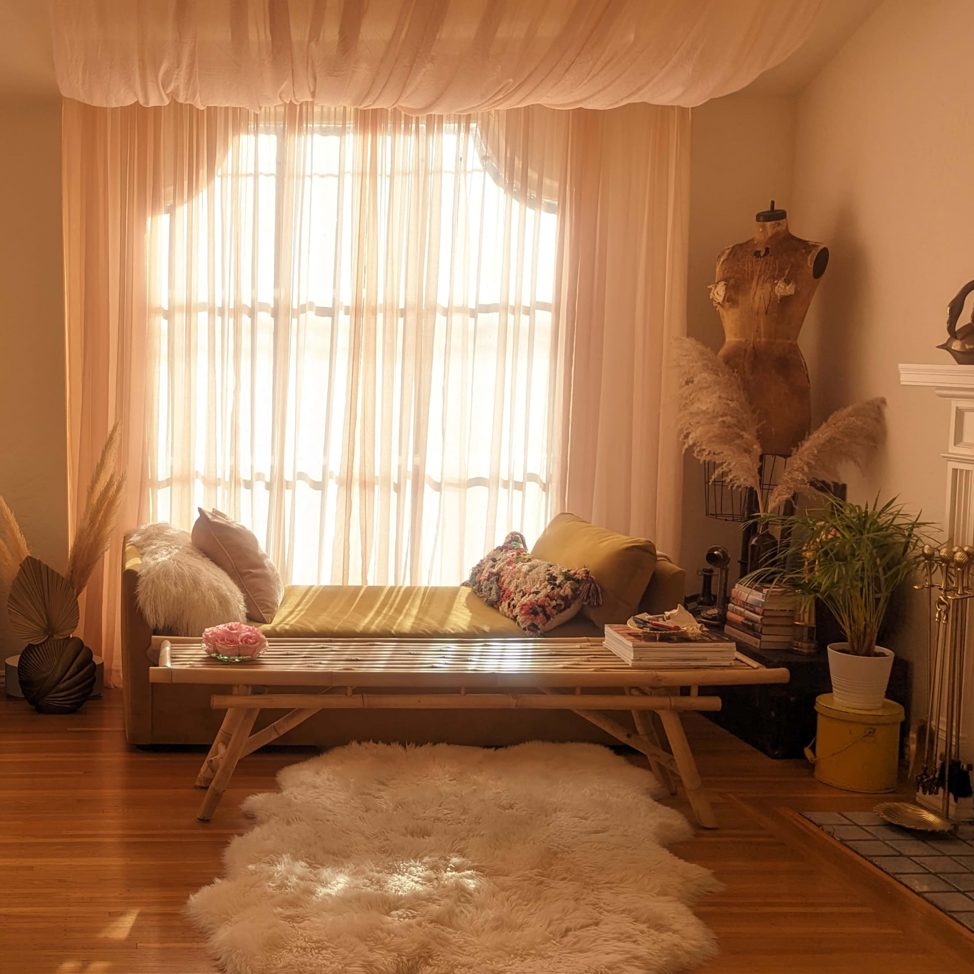 A softly lit room with large windows covered by sheer curtains, a wooden bench, a cozy seating area, and a decorative pole in the corner.