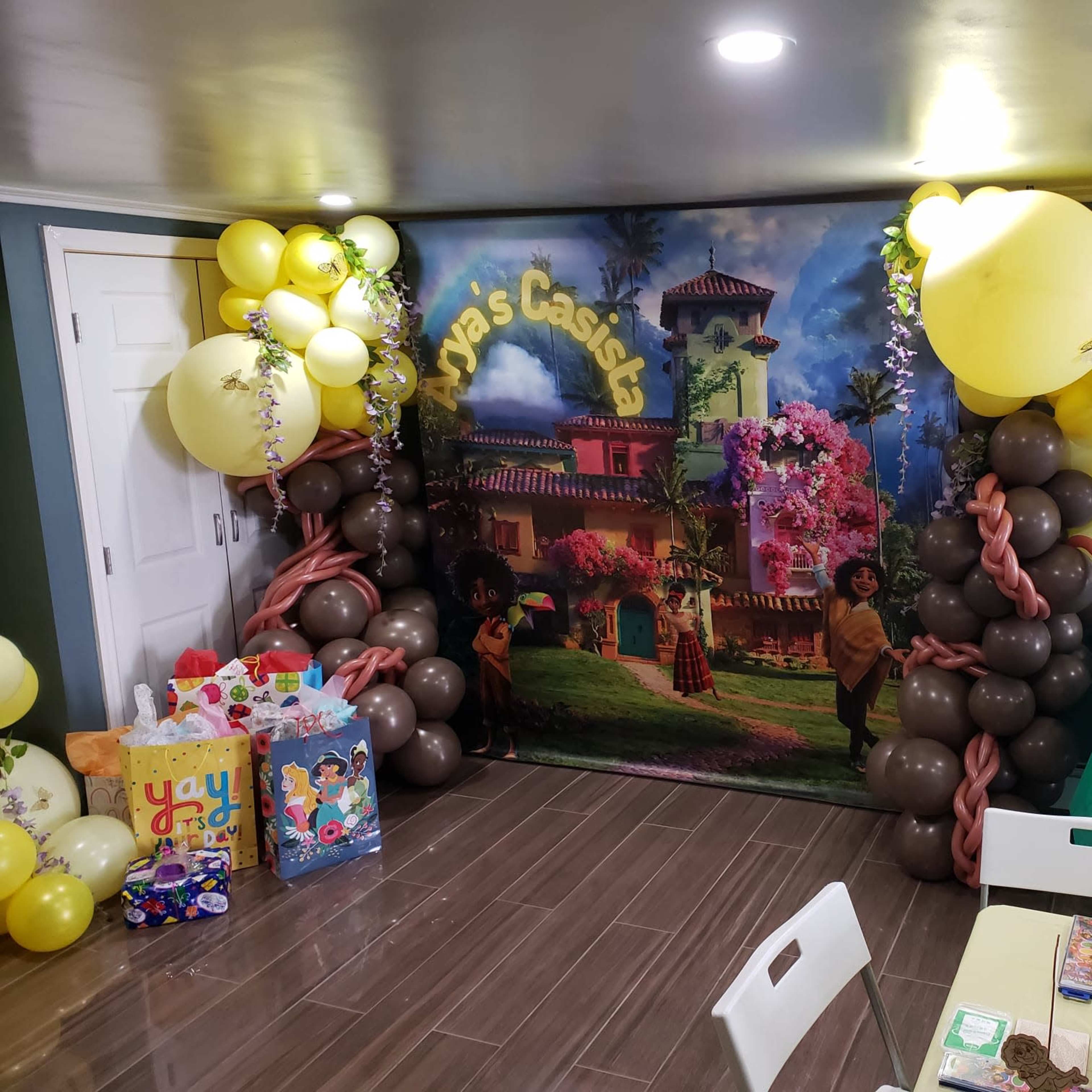 A decorated party room featuring a vibrant backdrop with a tropical theme, flanked by colorful balloons and gift bags.