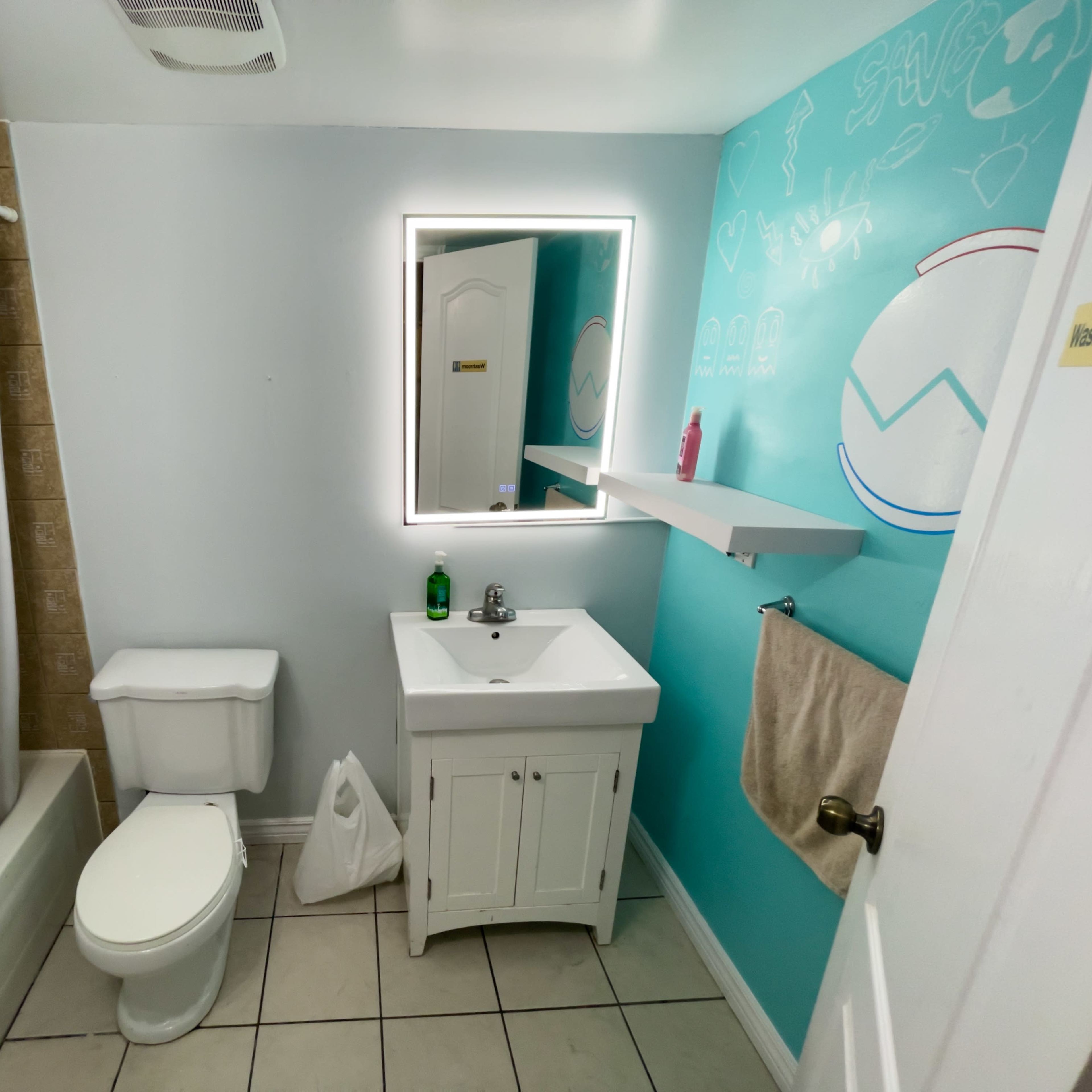 A bathroom features a shower/tub combination, a white vanity with a sink, a toilet, and a mirror with built-in lighting on a blue accent wall.
