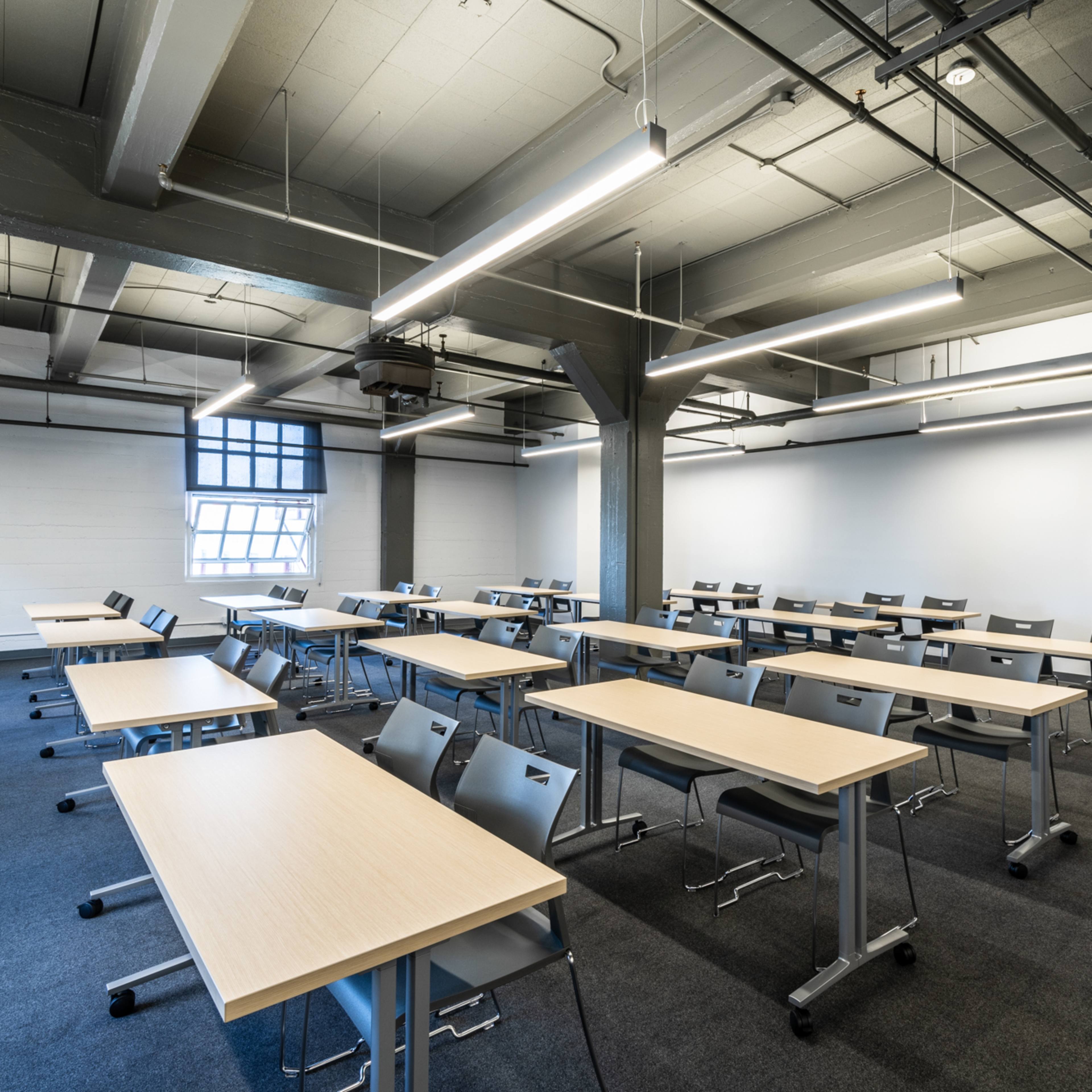 A classroom is filled with rows of empty desks and chairs, featuring industrial-style beams and overhead lighting.
