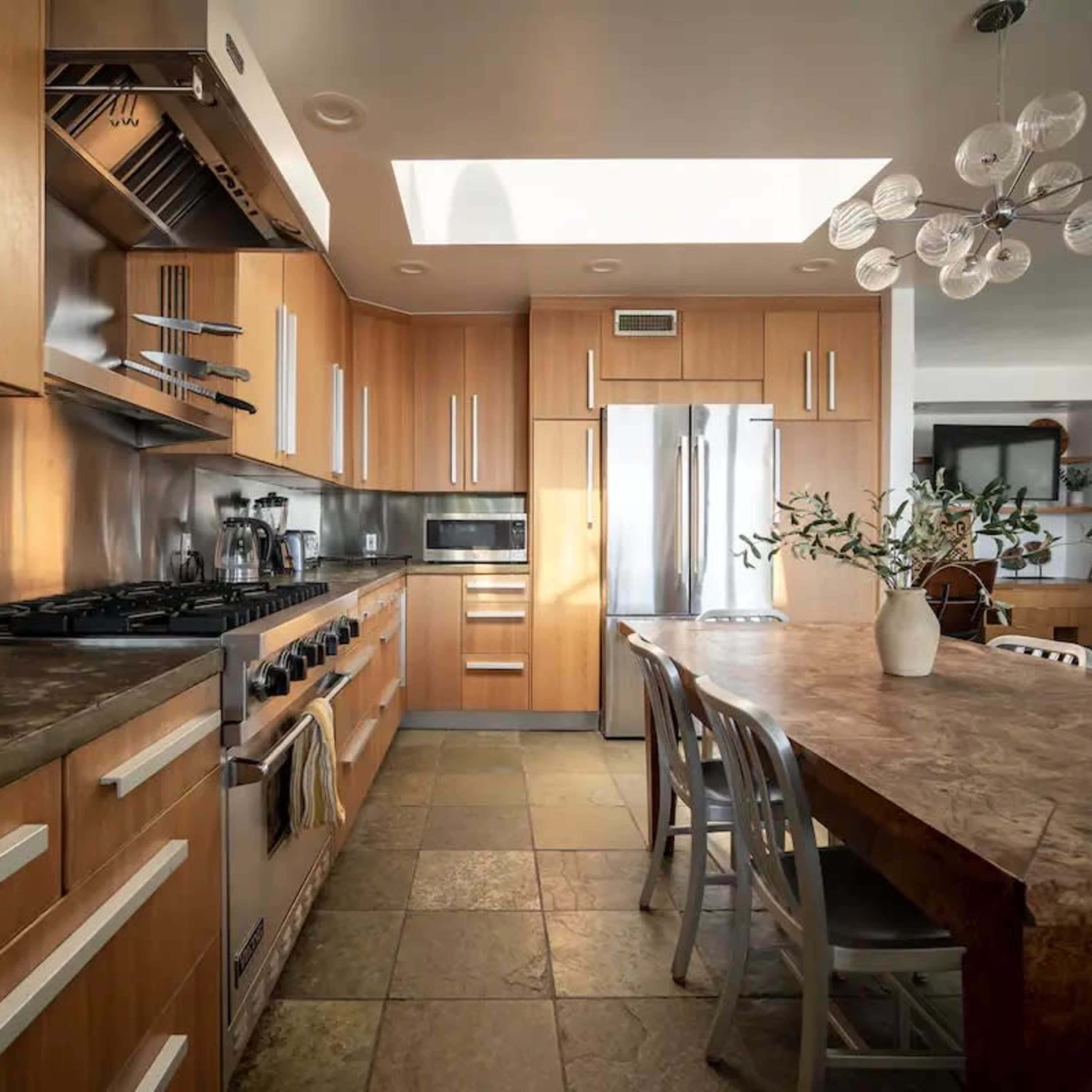 The kitchen features wooden cabinetry, stainless steel appliances, and a large dining table made of stone and wood.