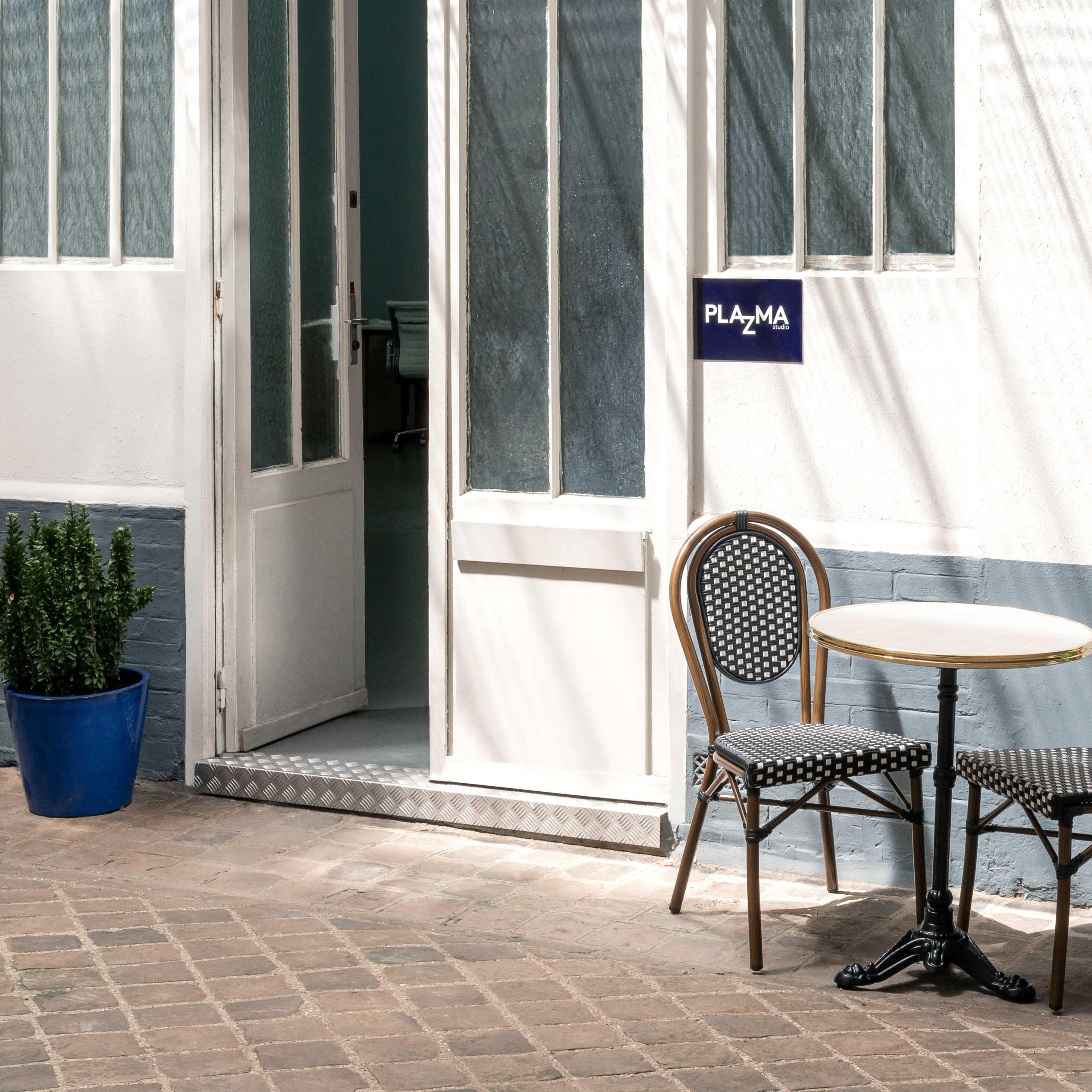 A small café table with two chairs is situated outside a building with a sign that reads "PLAZA," surrounded by potted plants.