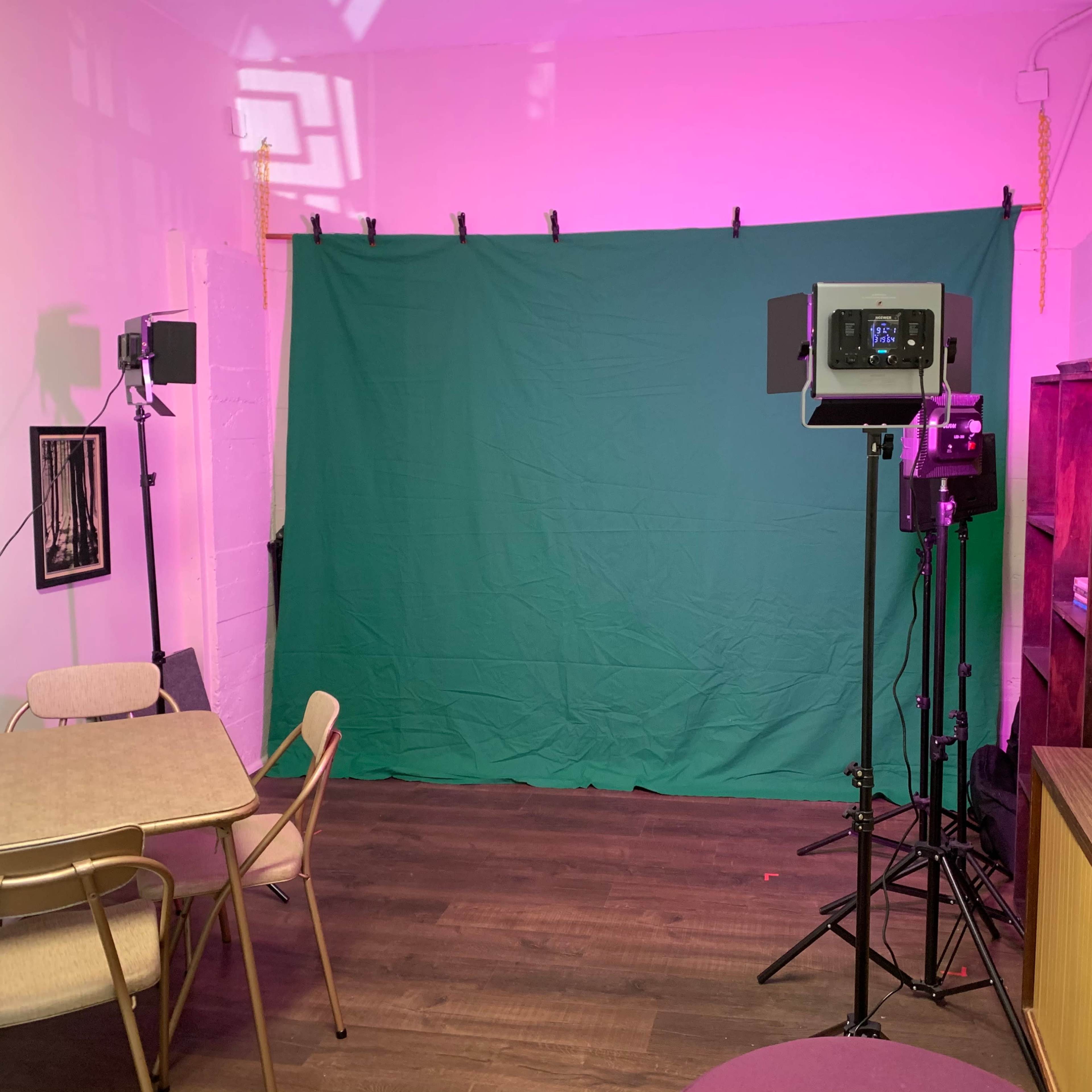 A studio space features a green backdrop, two lights, a purple couch, a coffee table, and a rug on the wooden floor.