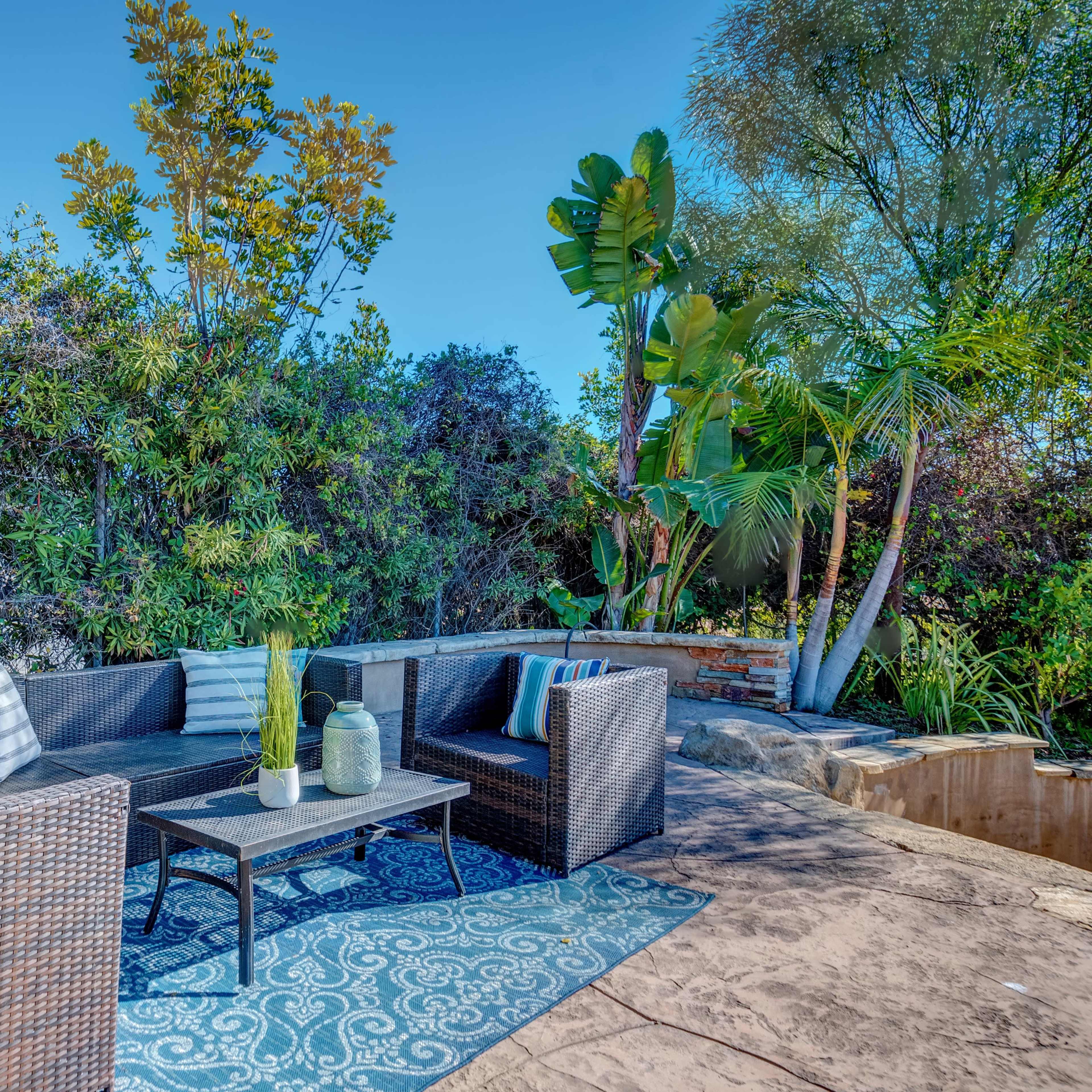 A cozy outdoor seating area features a black wicker sofa set arranged around a coffee table on a patterned rug, surrounded by lush greenery.