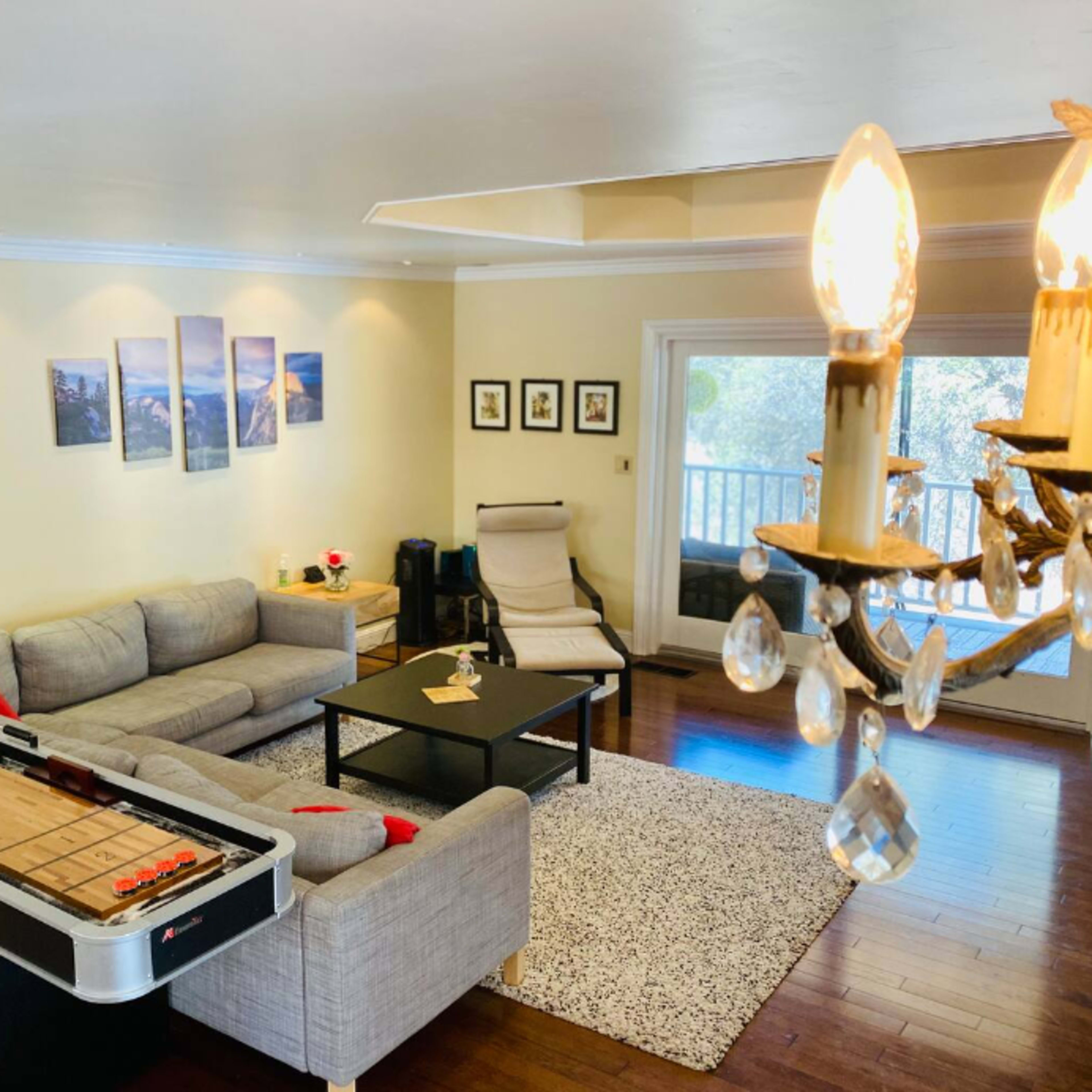 The room features a gray sectional sofa, a coffee table, a shuffleboard table, and large windows overlooking a balcony.