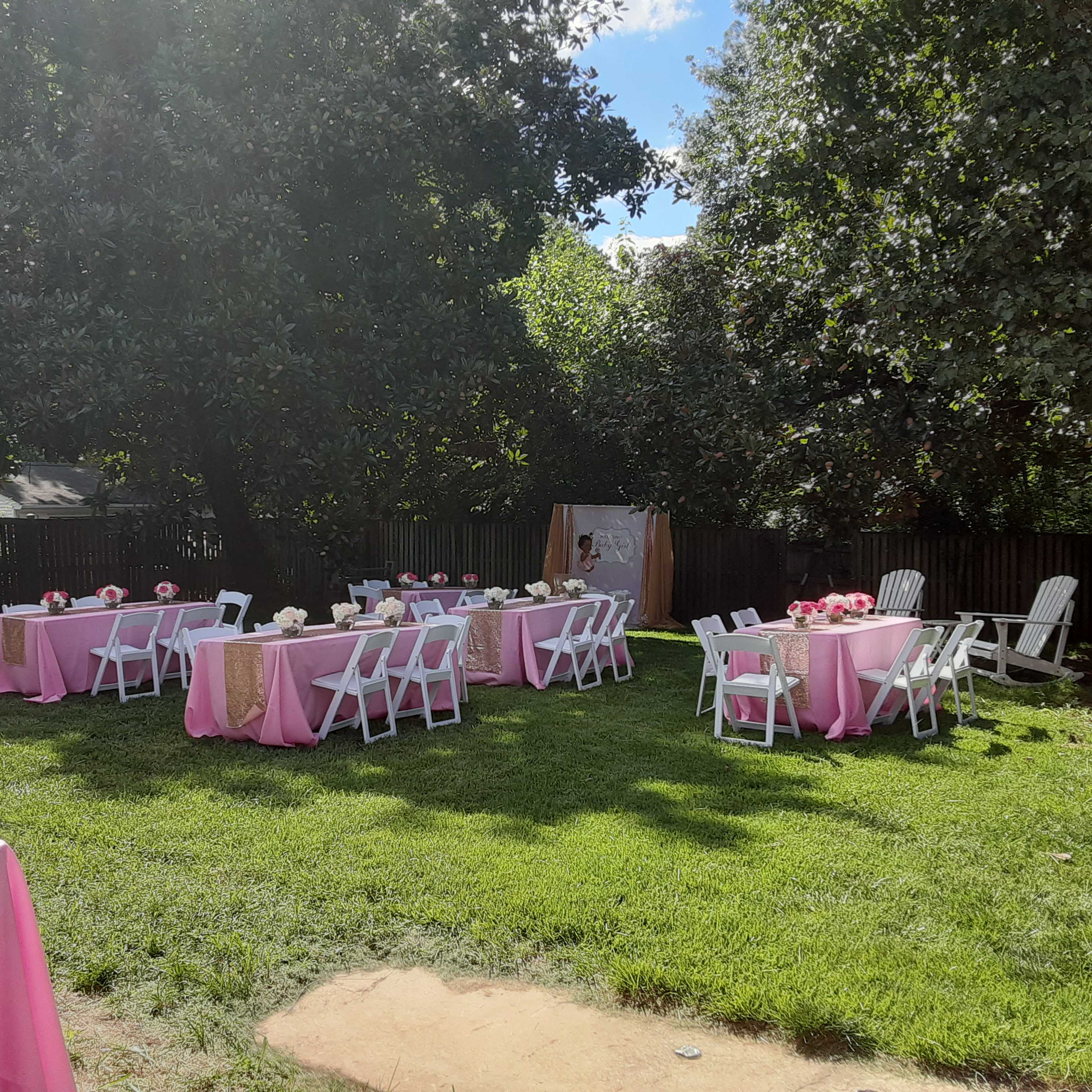 A backyard set up for an outdoor event, featuring several tables covered with pink tablecloths and surrounded by white chairs, all situated on green grass.