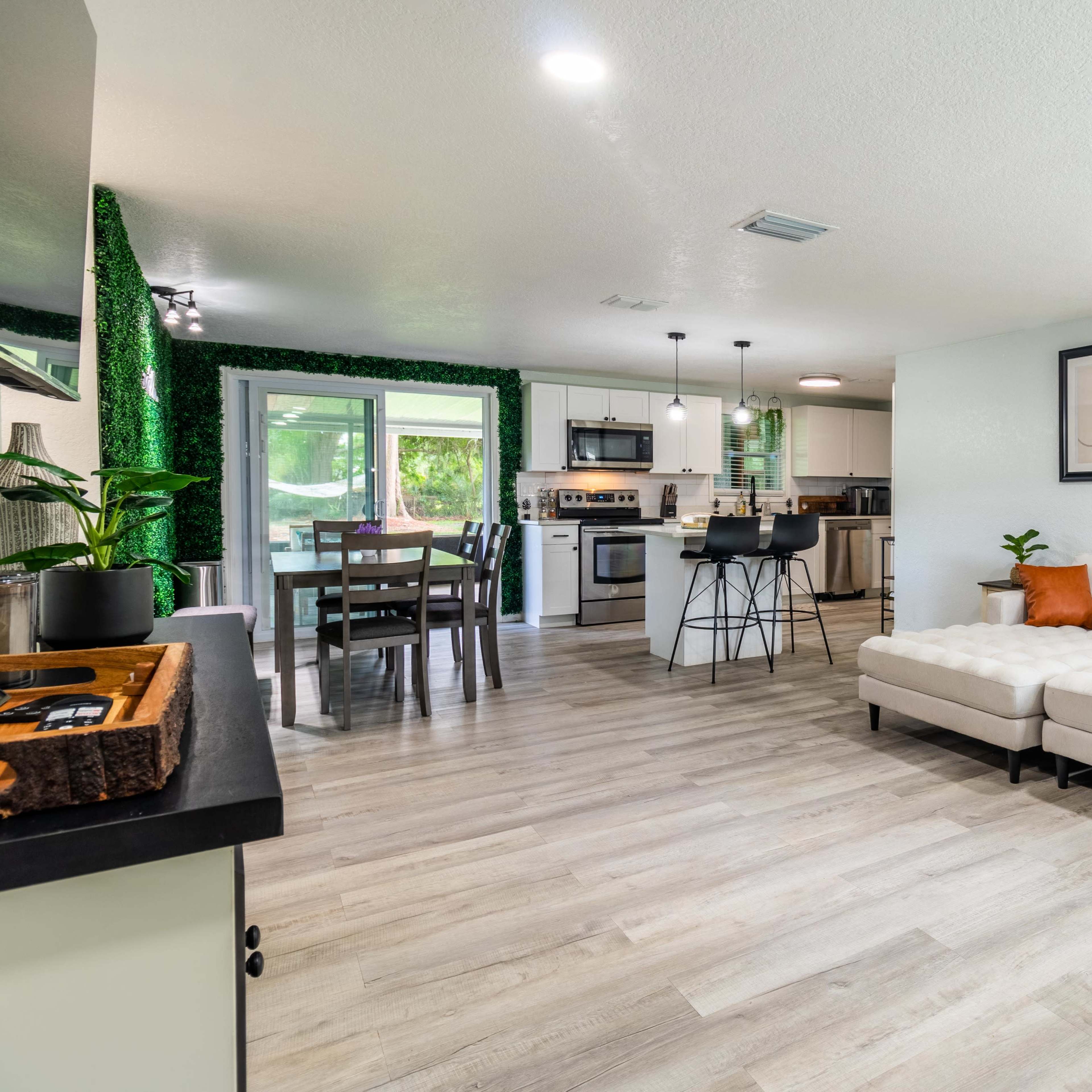 A modern living space with a white sectional sofa, a dining area, and a kitchen, all featuring light wood flooring and contemporary decor.
