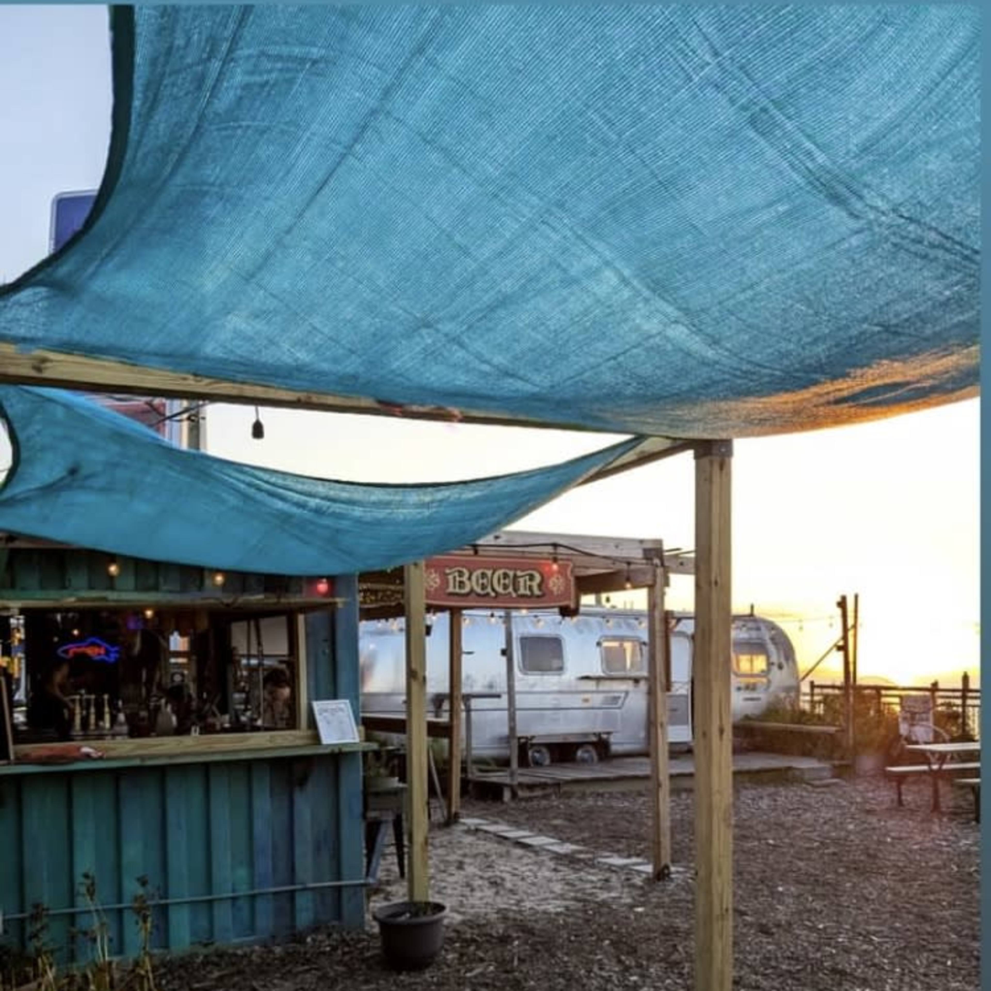 The scene shows a bar with a blue canopy and vintage trailers in the background, set along a waterfront area during sunset.