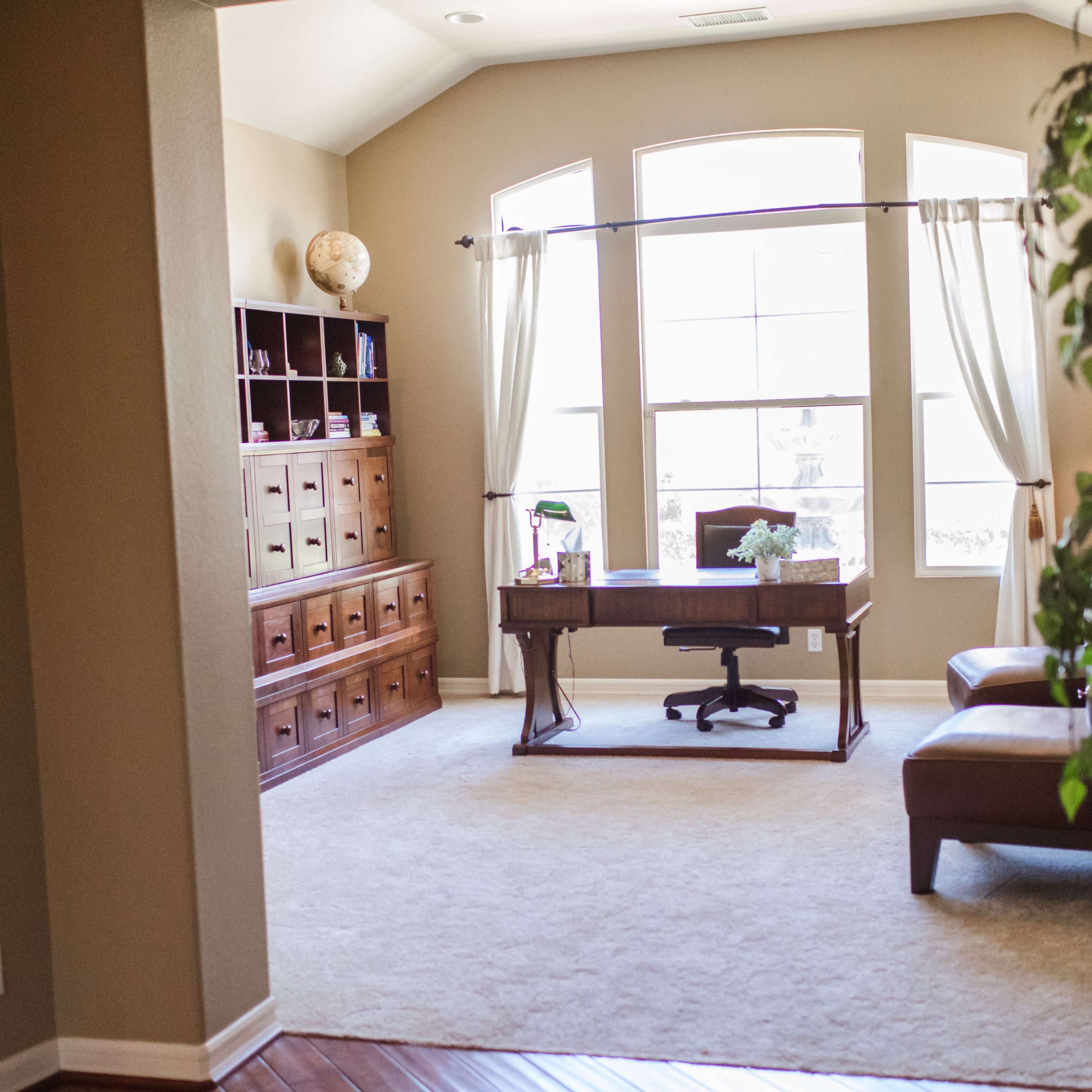 A home office featuring a wooden desk with an ergonomic chair, a large window with sheer curtains, a shelving unit, and a potted plant in the corner.