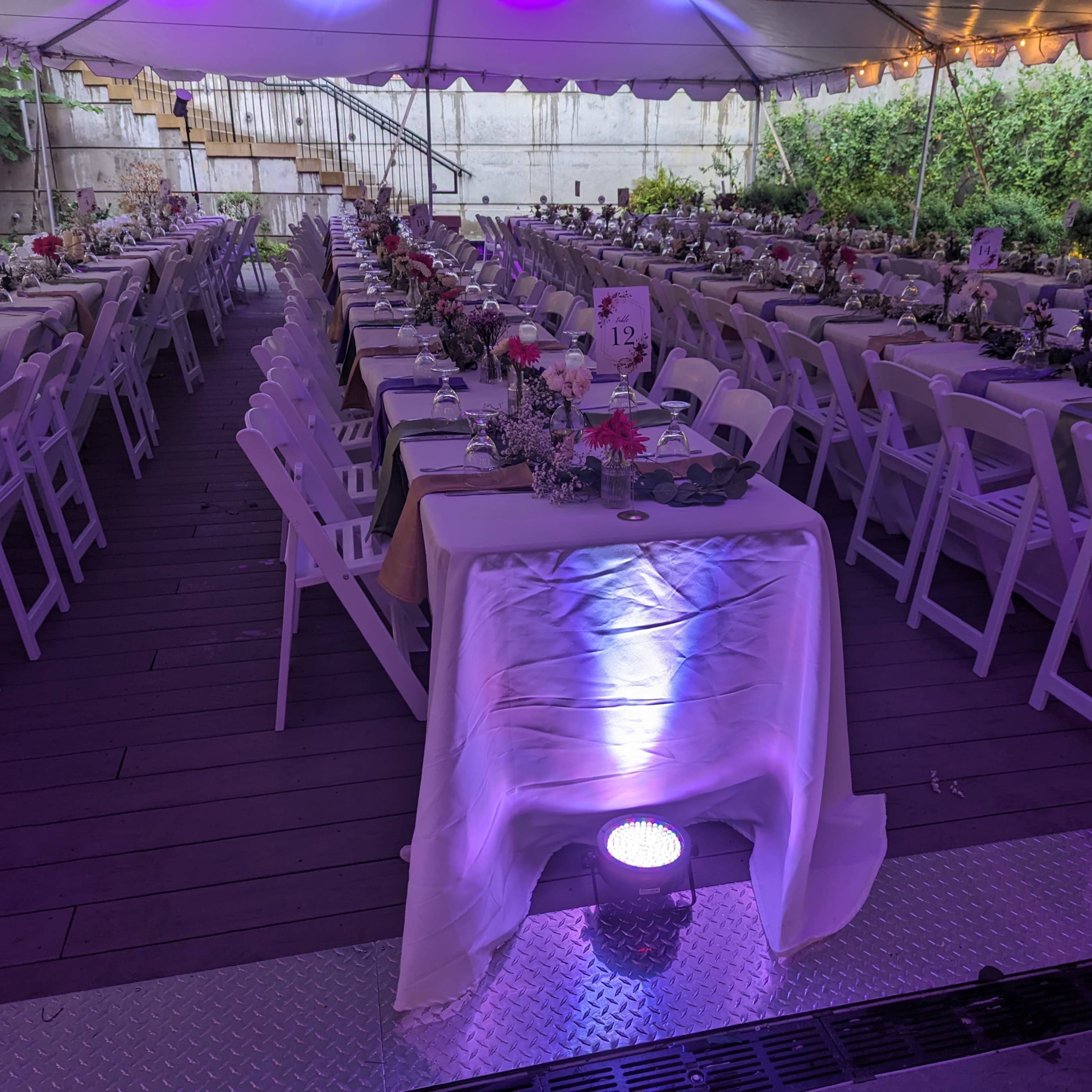 A large tented area is set up for a dining event, featuring rows of white tables adorned with floral centerpieces and surrounded by white chairs.