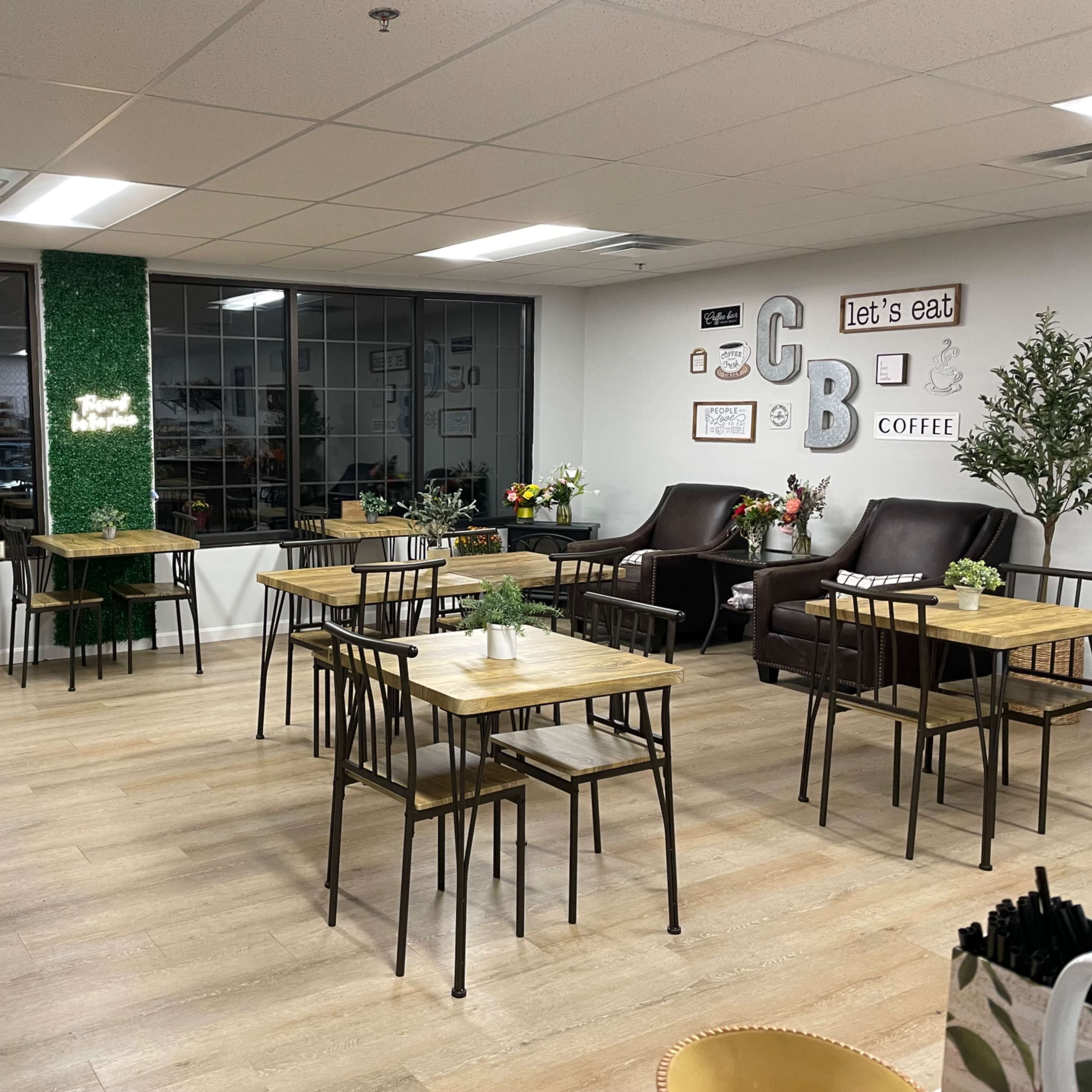 A cozy café interior with several wooden tables and chairs, large windows, and decorative plants.