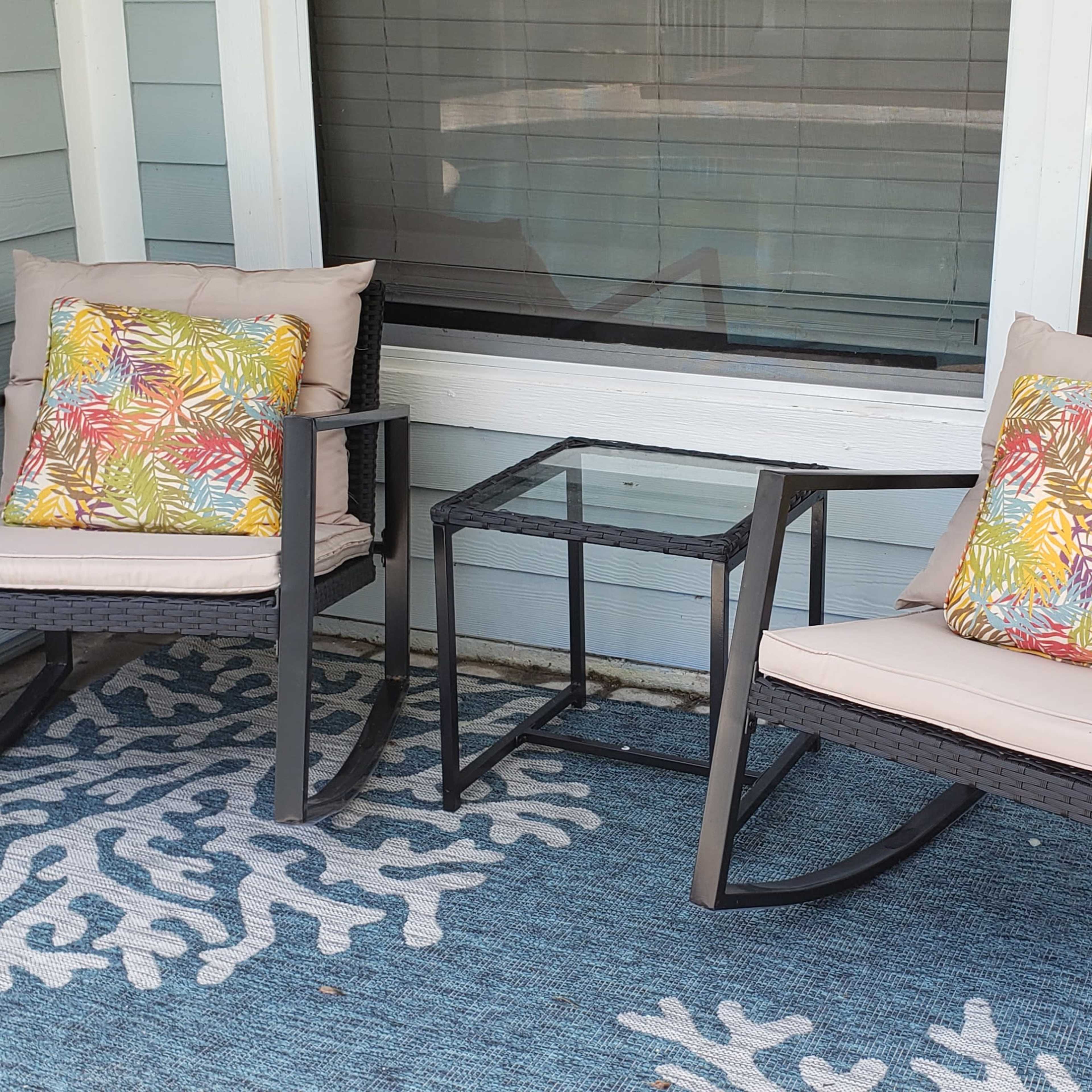 Two rocking chairs with colorful cushions sit beside a glass table on a patterned rug in a porch area.