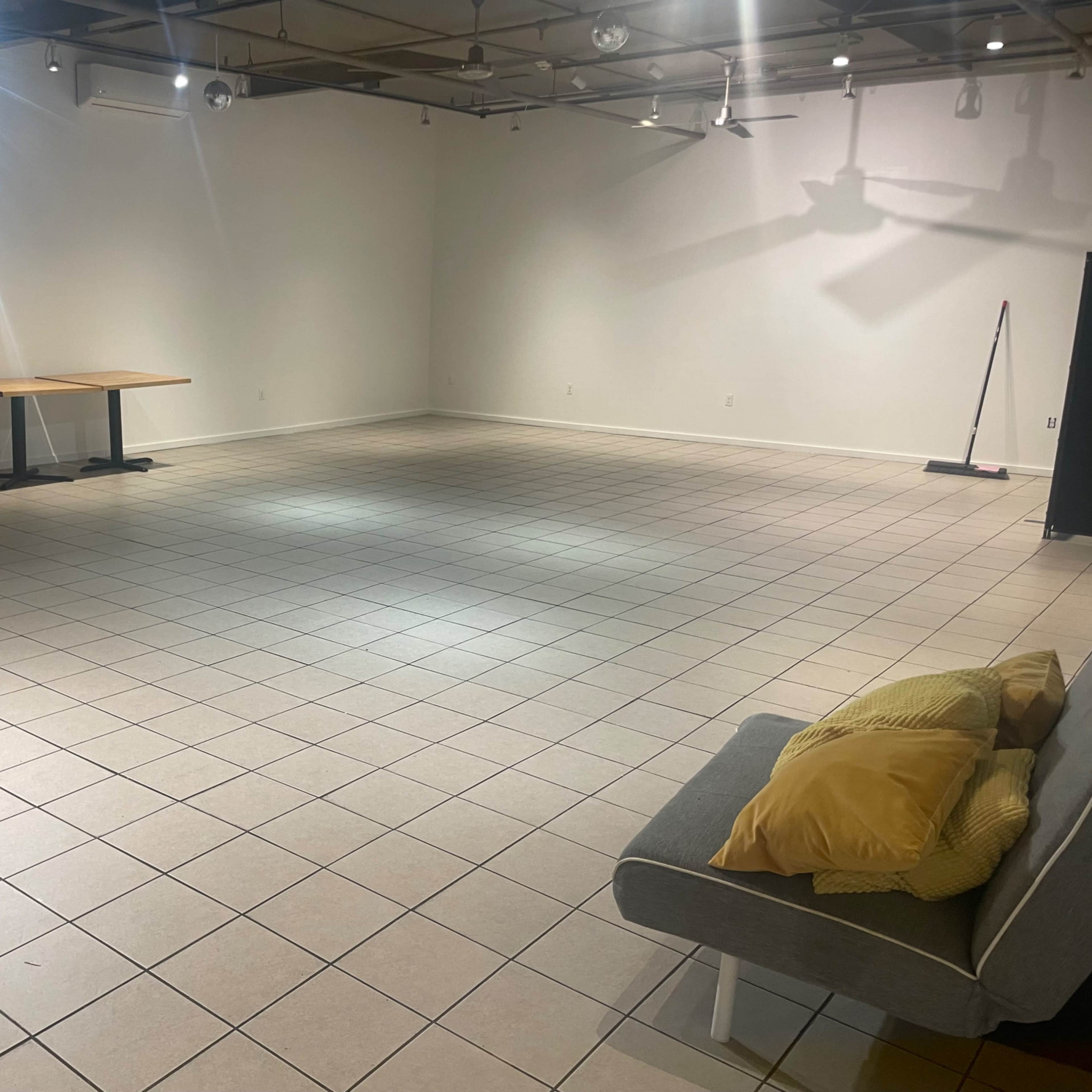 A spacious, empty room with tiled flooring, some tables lined against the wall, and a sofa with yellow cushions in one corner.