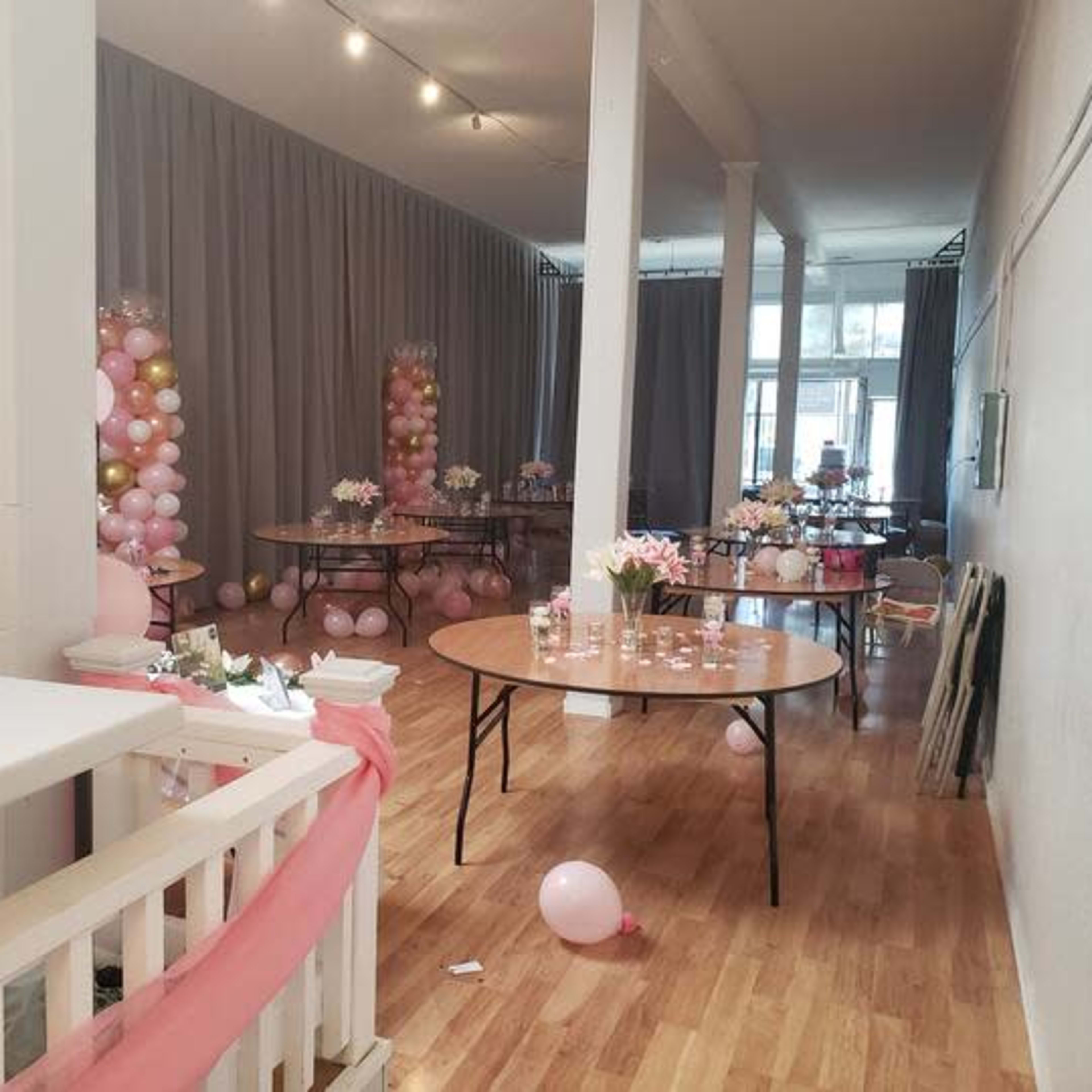 A decorated event space with round tables, floral centerpieces, and balloon arrangements in pink and gold.