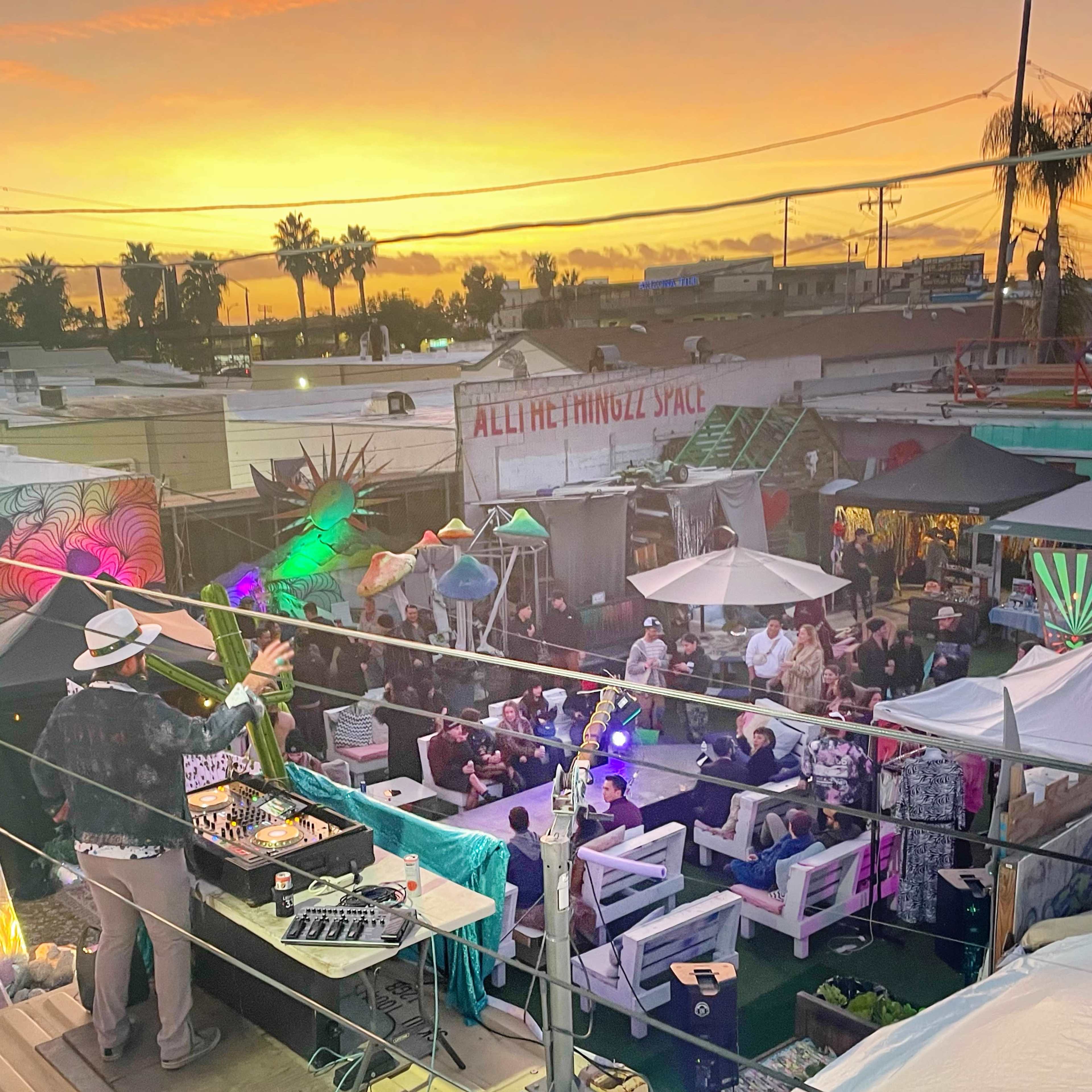 A DJ performs on an elevated stage while a crowd gathers in a vibrant outdoor space at sunset.