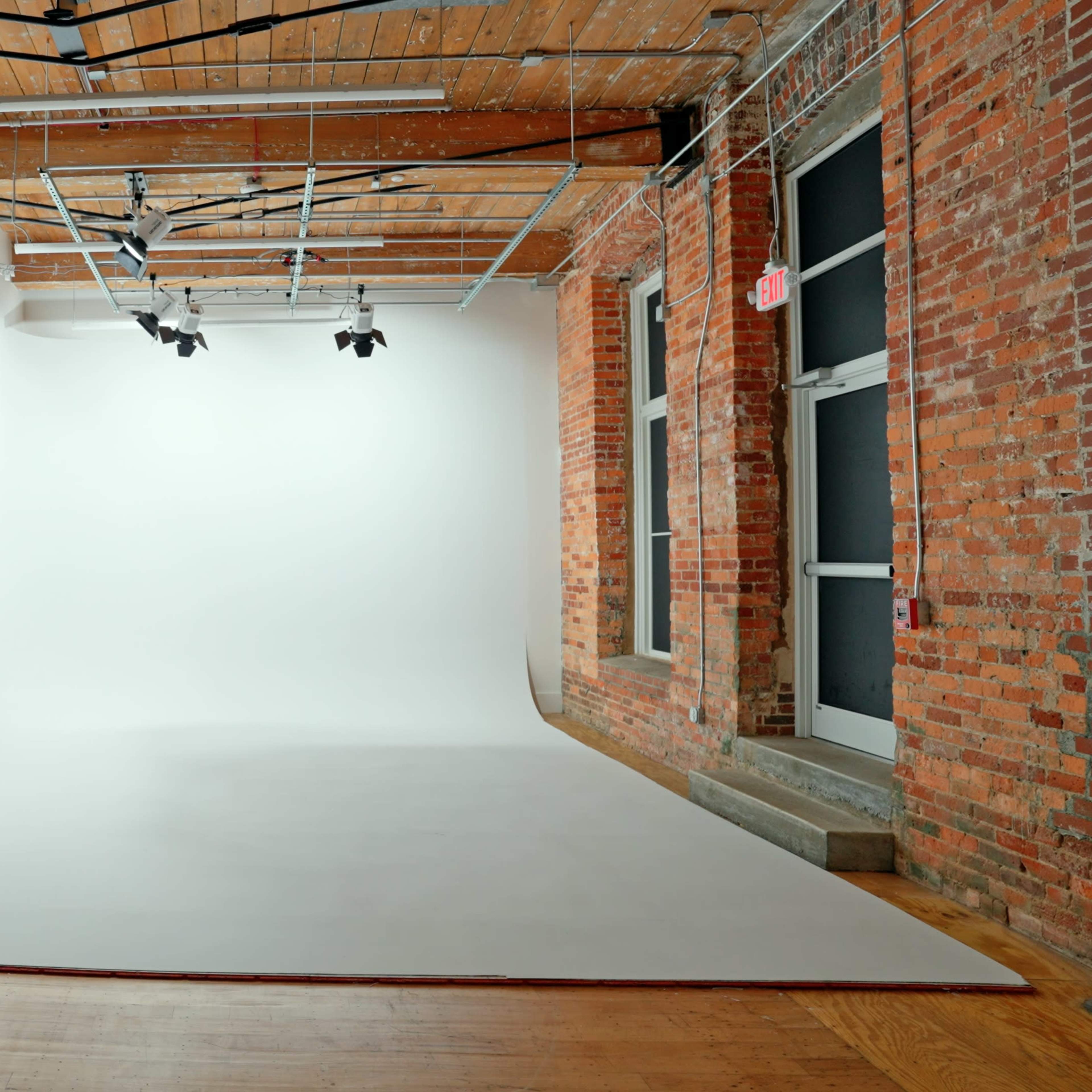 A spacious photography studio with a large white backdrop, wooden floor, and exposed brick walls.