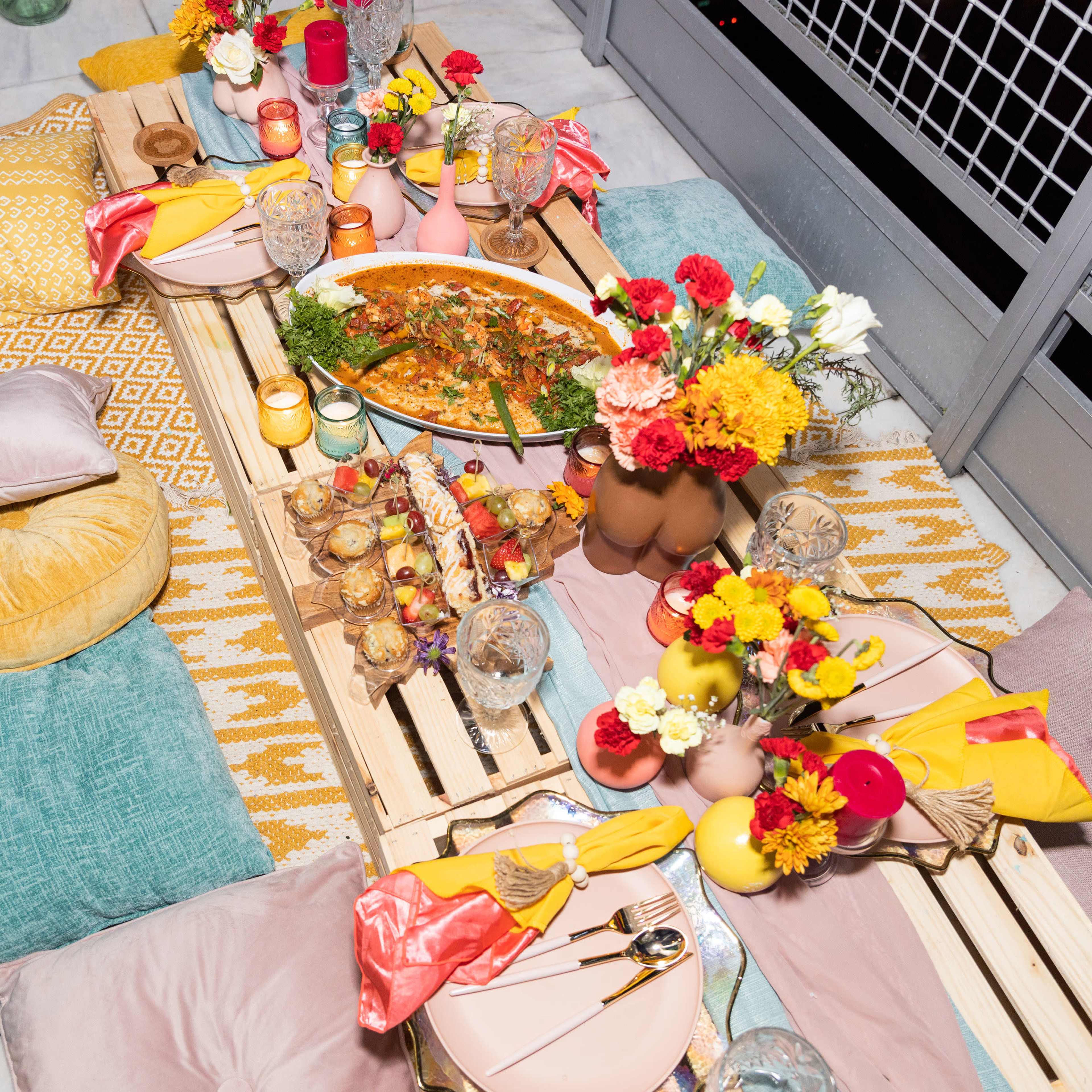 A vibrant outdoor dining setup features a long table adorned with colorful dishes, glassware, and cushions on a balcony.