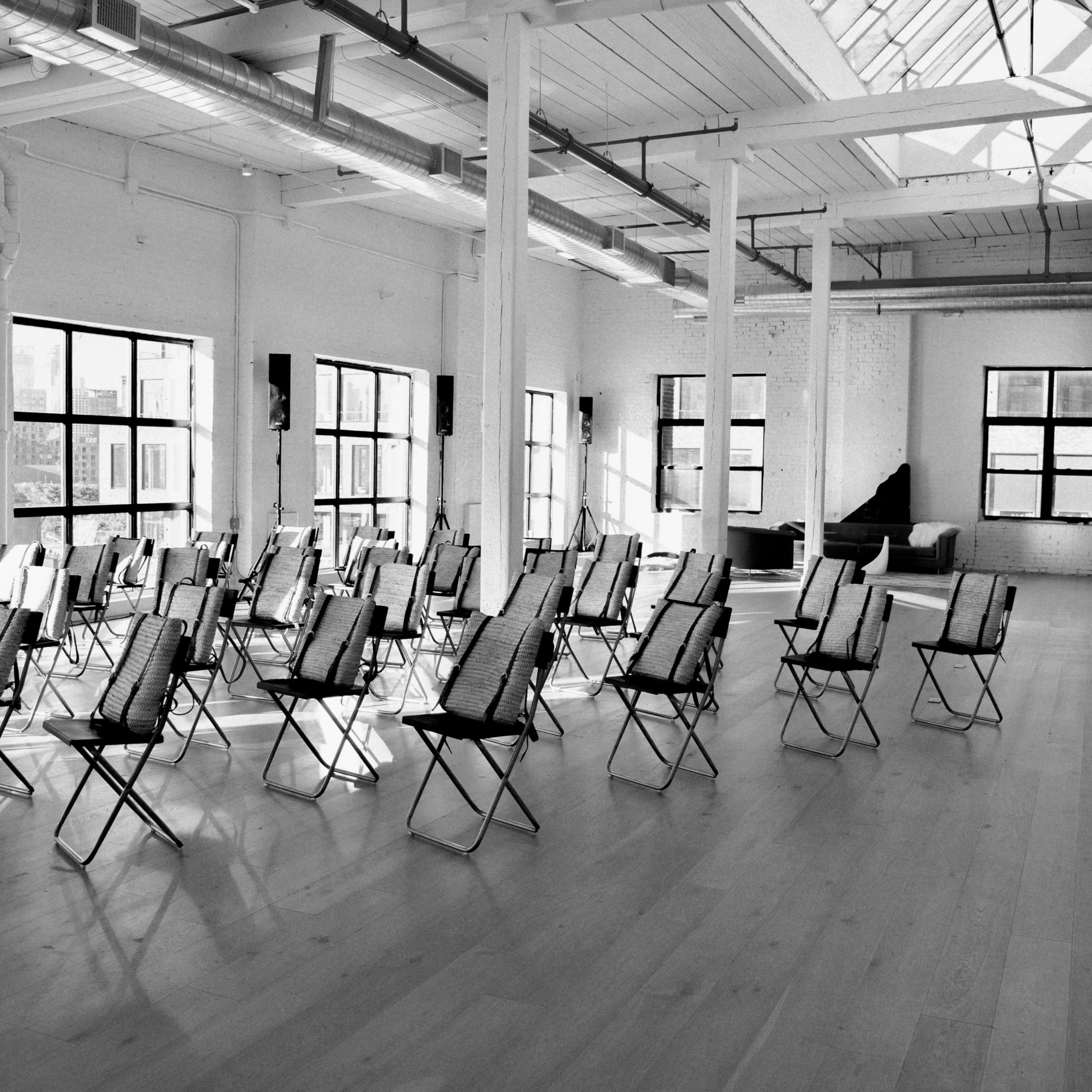 A spacious, brightly lit room arranged with multiple rows of folding chairs facing a central area, with large windows providing natural light.
