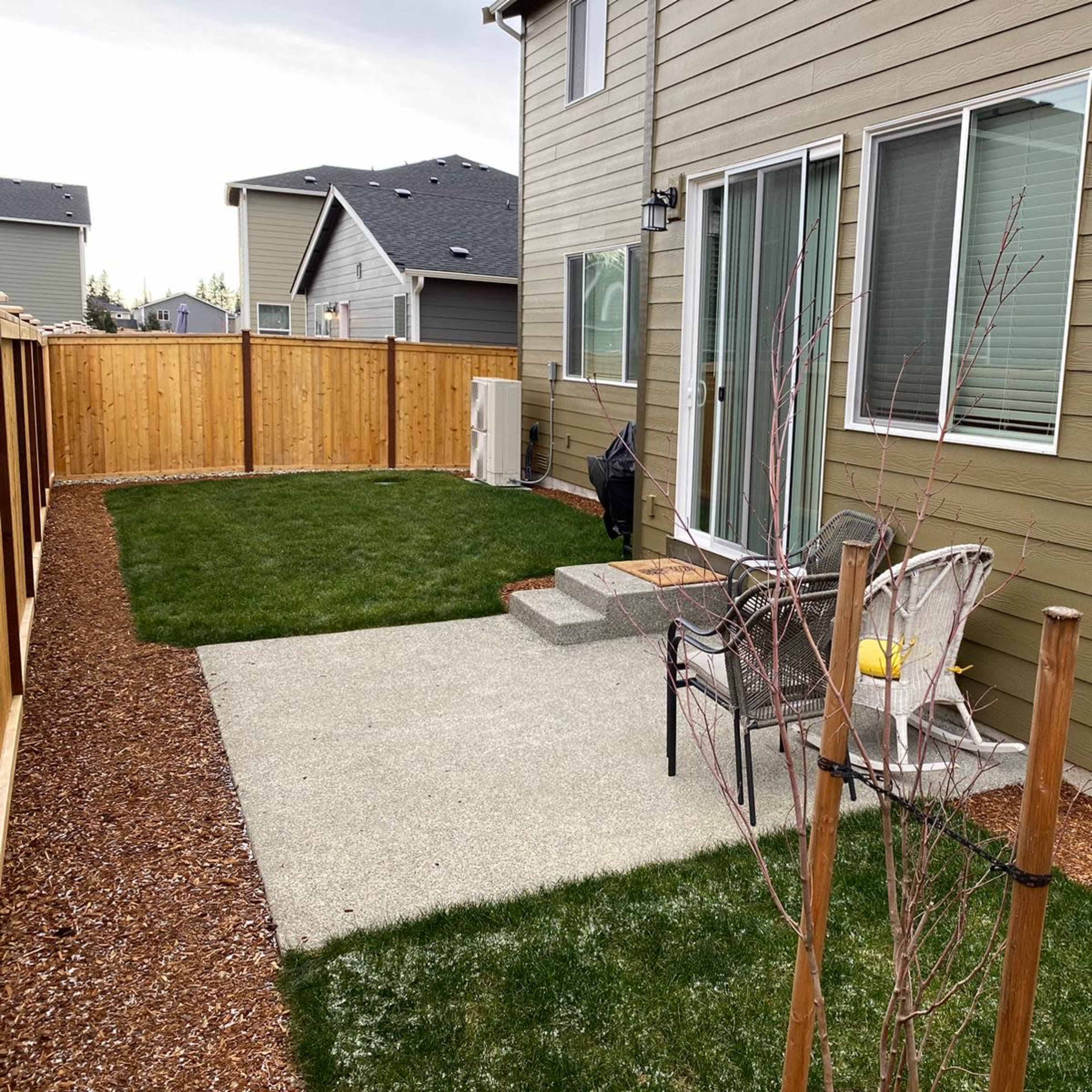 A neatly landscaped backyard with a concrete patio, grassy areas, and a small tree near a wooden fence.