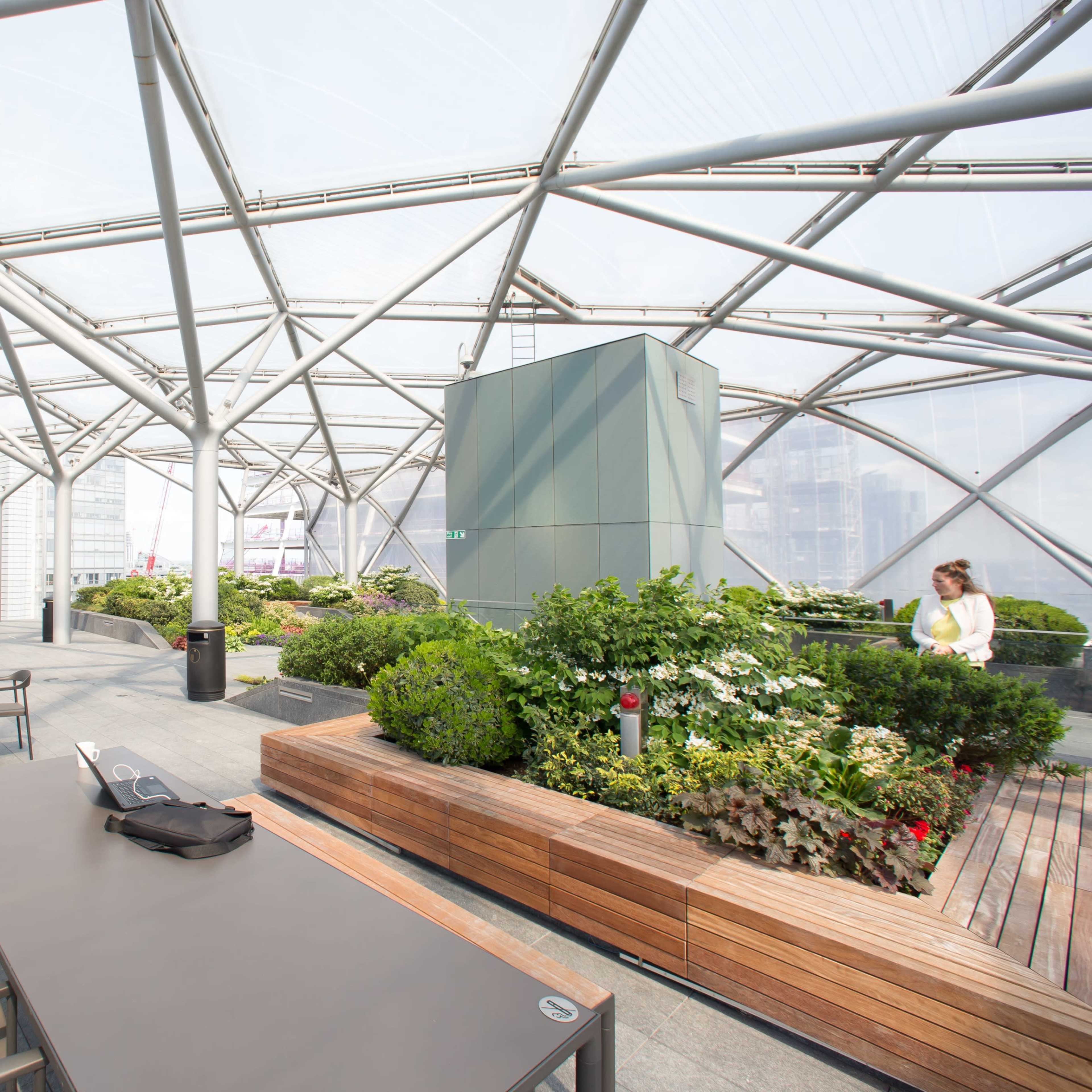 A modern rooftop terrace with metal framework, green plants, and wooden seating areas overlooking a cityscape.