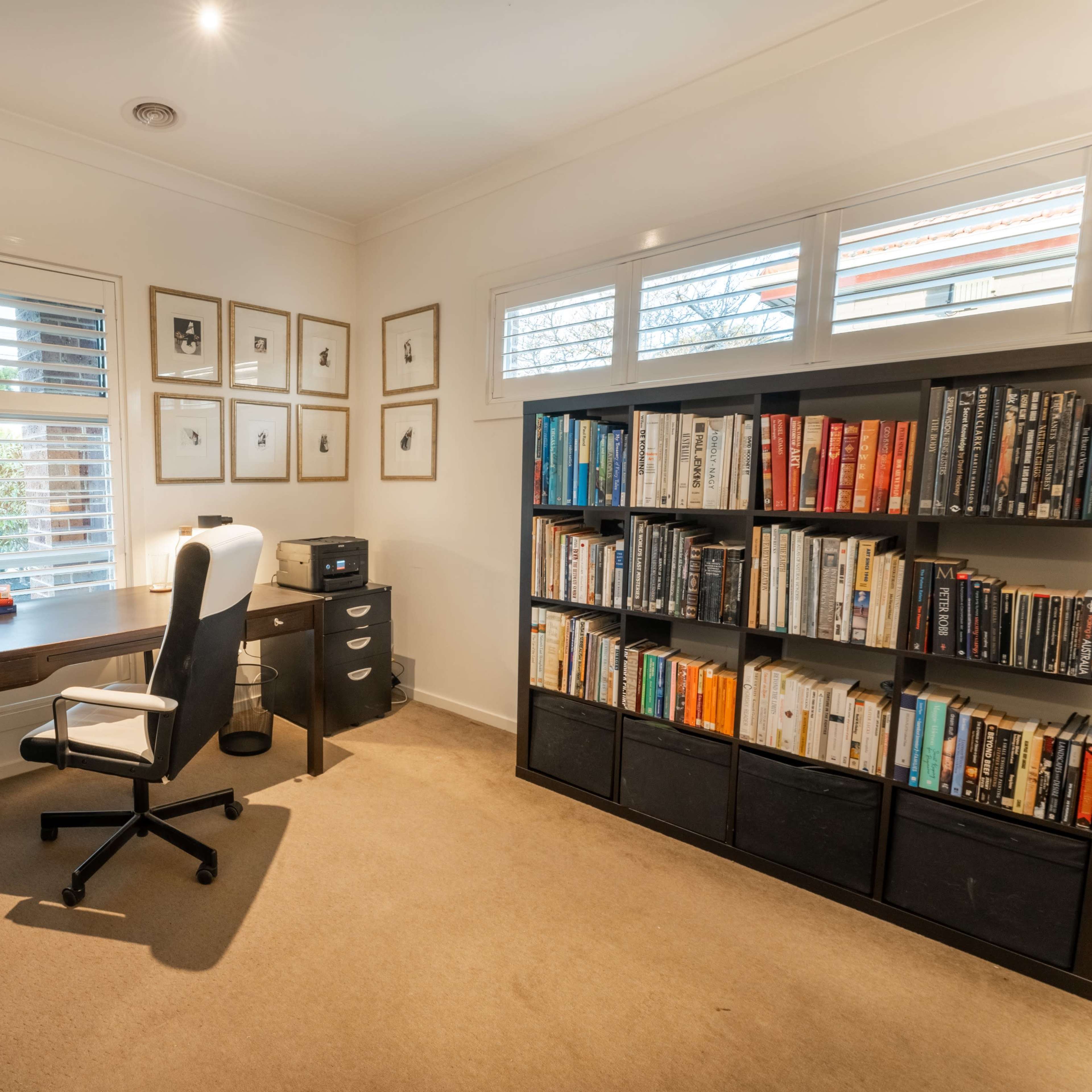 A tidy home office features a desk with a computer monitor, a swivel chair, a bookshelf filled with books, and framed artwork on the walls.