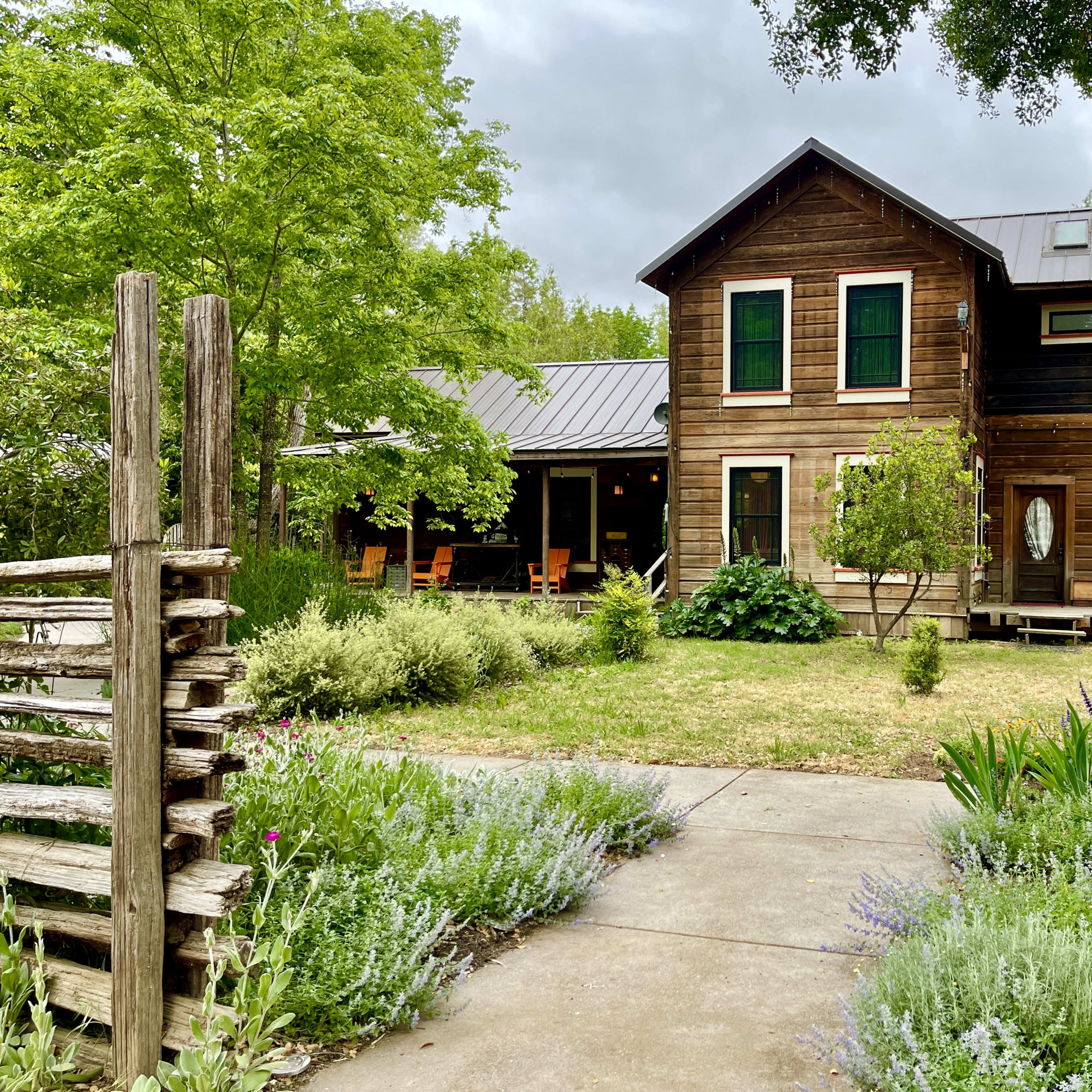 A rustic wooden house with a metal roof is surrounded by greenery and a gravel pathway.