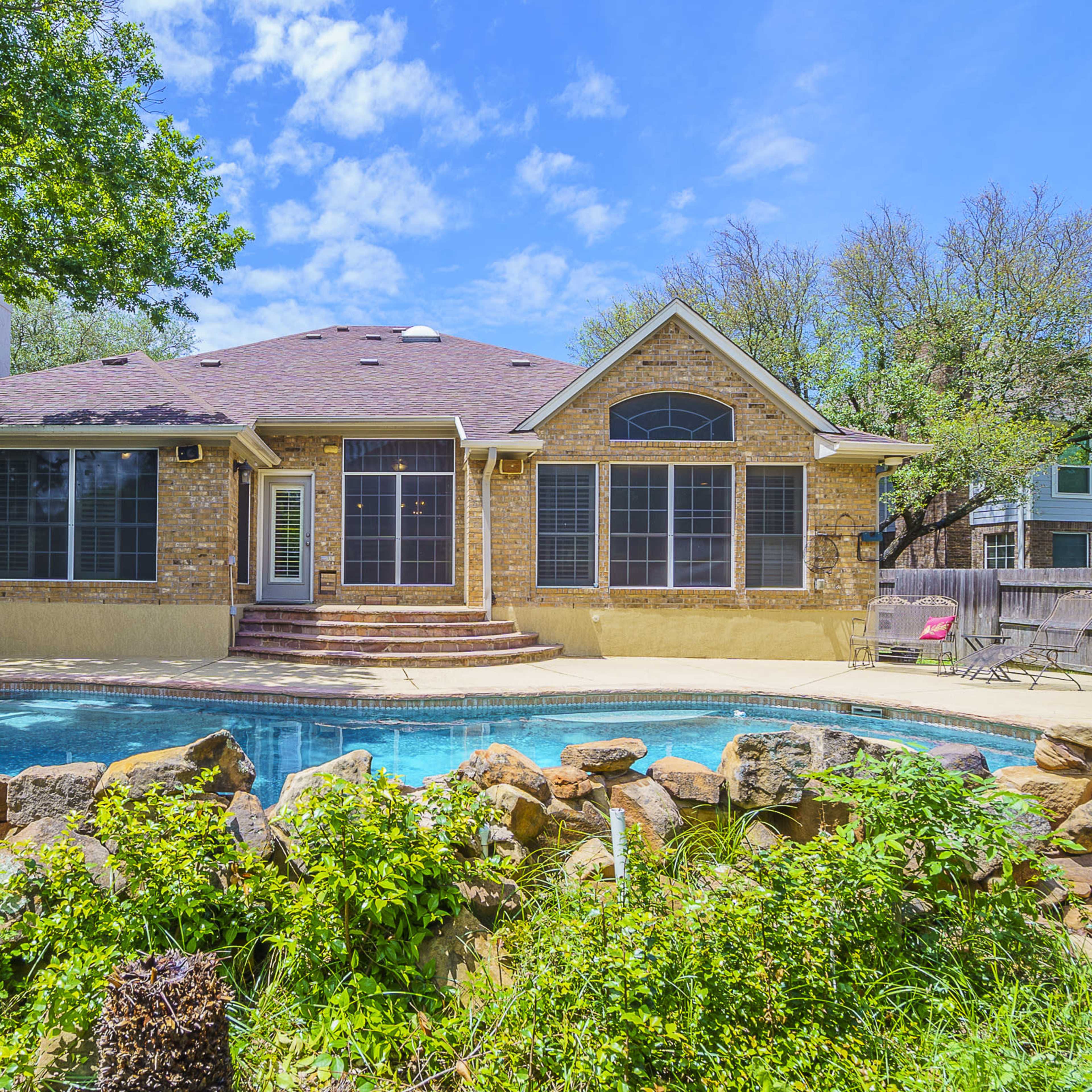 A single-story stone house with large windows overlooks a backyard pool surrounded by a stone wall and lush greenery.