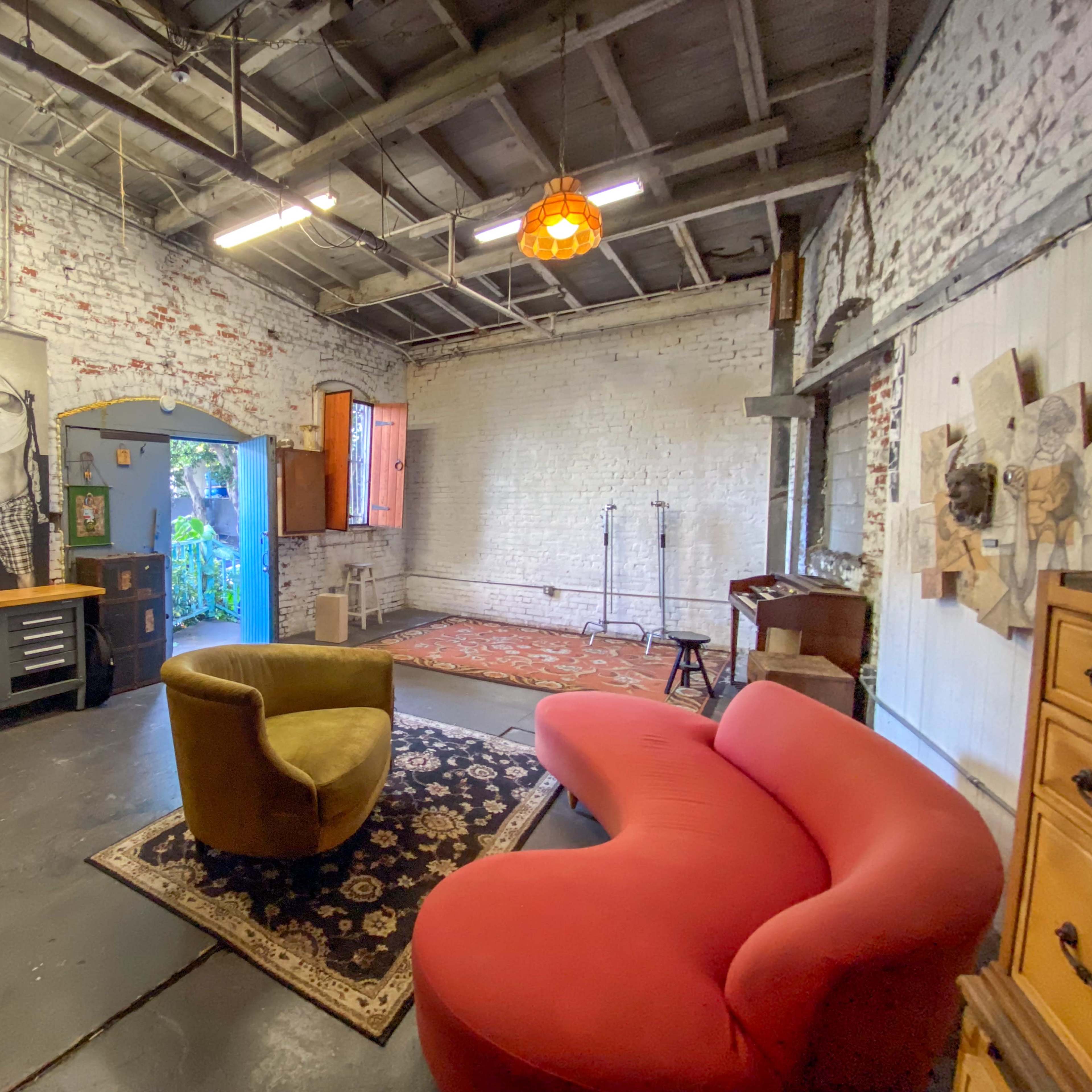 A spacious room with exposed brick walls, featuring a green and red modern couch, a patterned rug, and various furniture pieces scattered throughout.