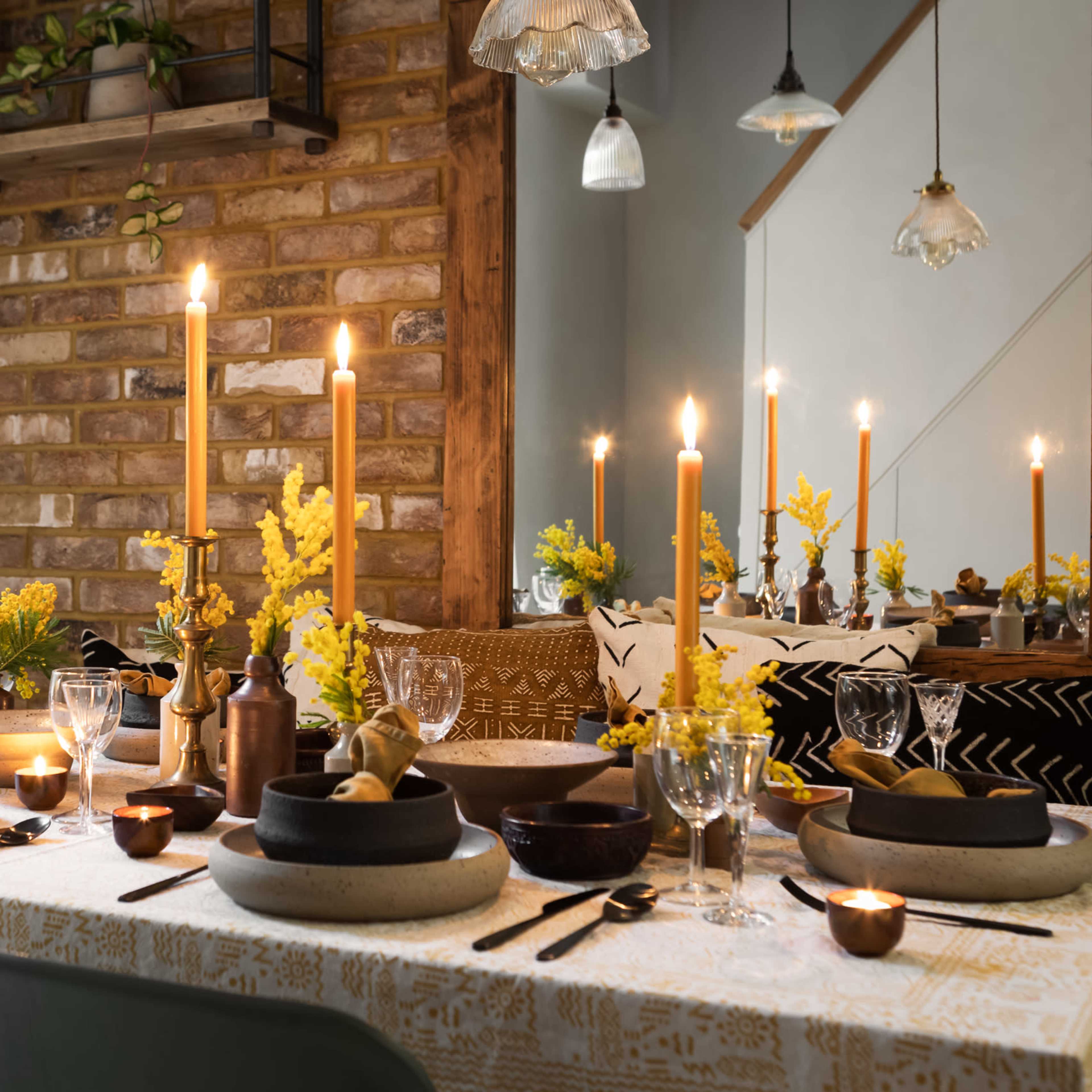 A dining table is set with candles, flowers, and various tableware against a backdrop of exposed brick and decorative lighting.