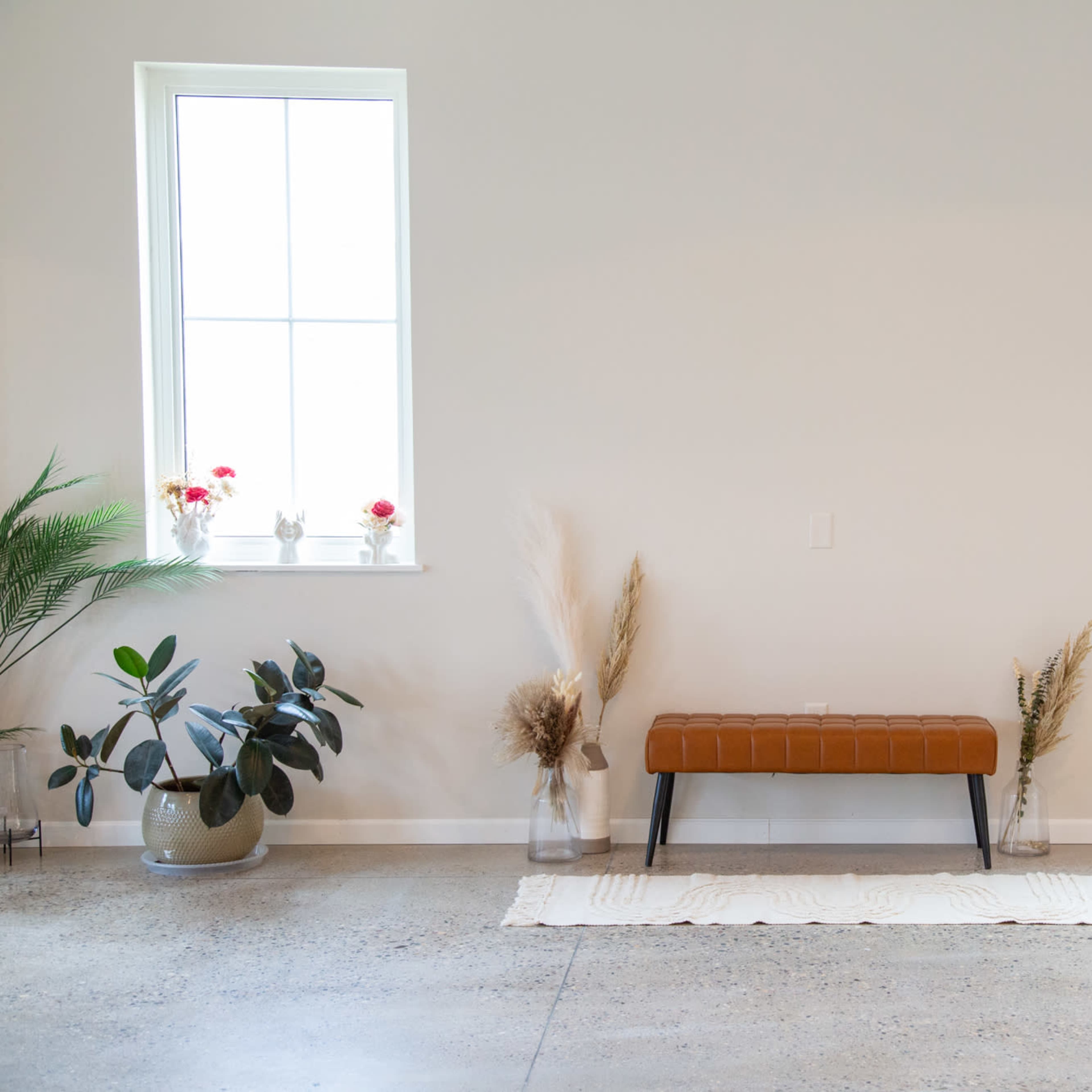 A minimalist interior with a brown bench centered against a light-colored wall, flanked by plants and decorative elements near two windows.