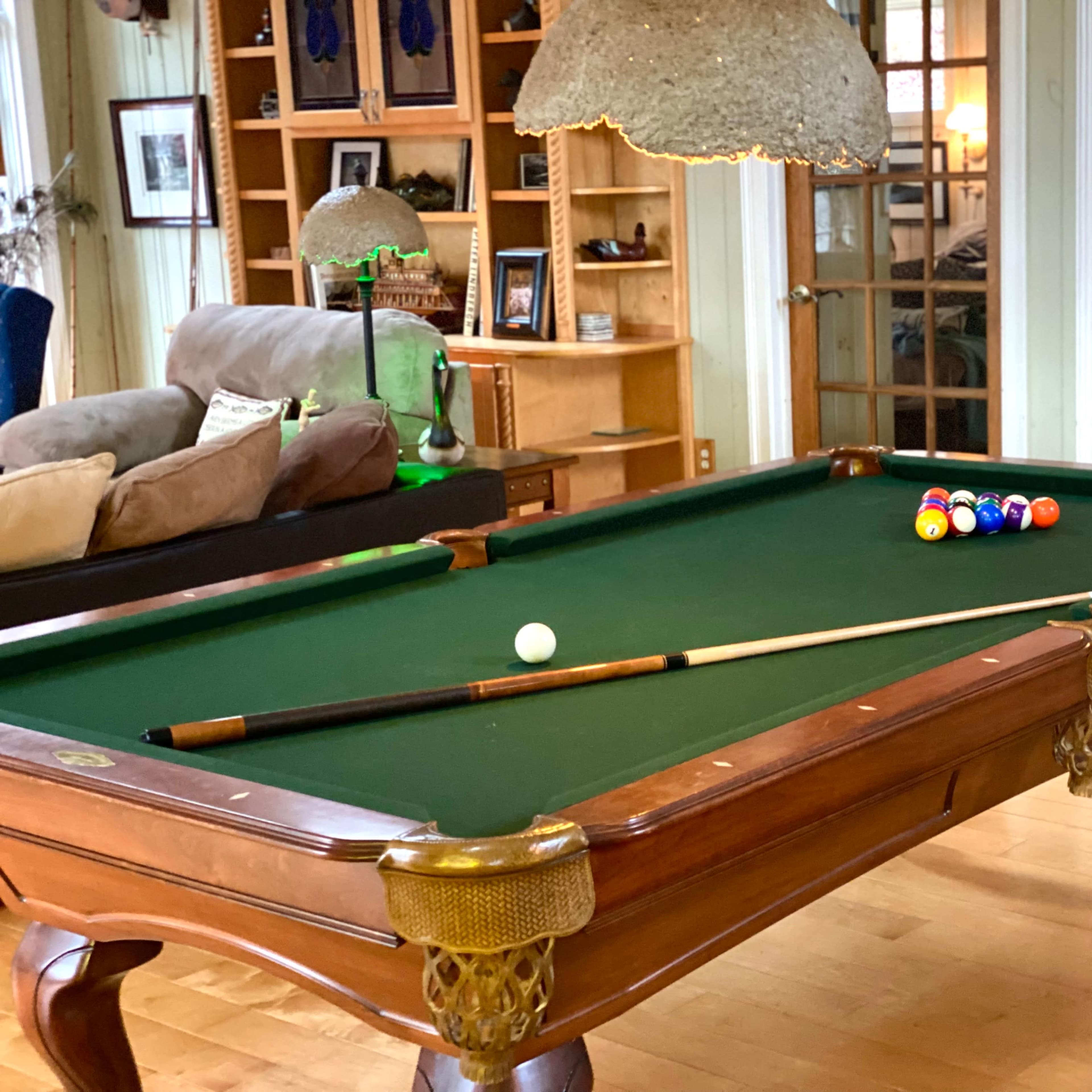 A green-felt billiard table with a cue stick and a set of colored balls arranged on its surface, located in a well-furnished room.