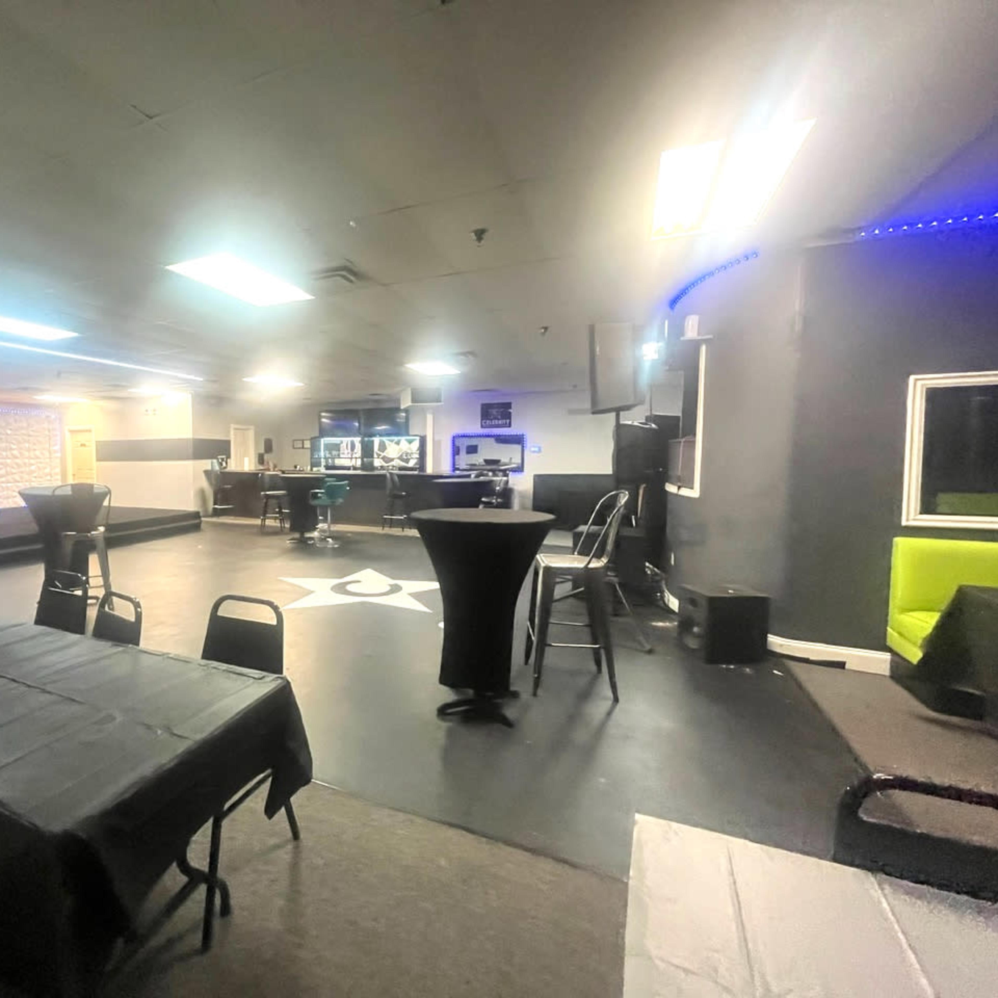 An empty, dimly lit event space with tables and chairs arranged around a central dance floor and a bar at one end.