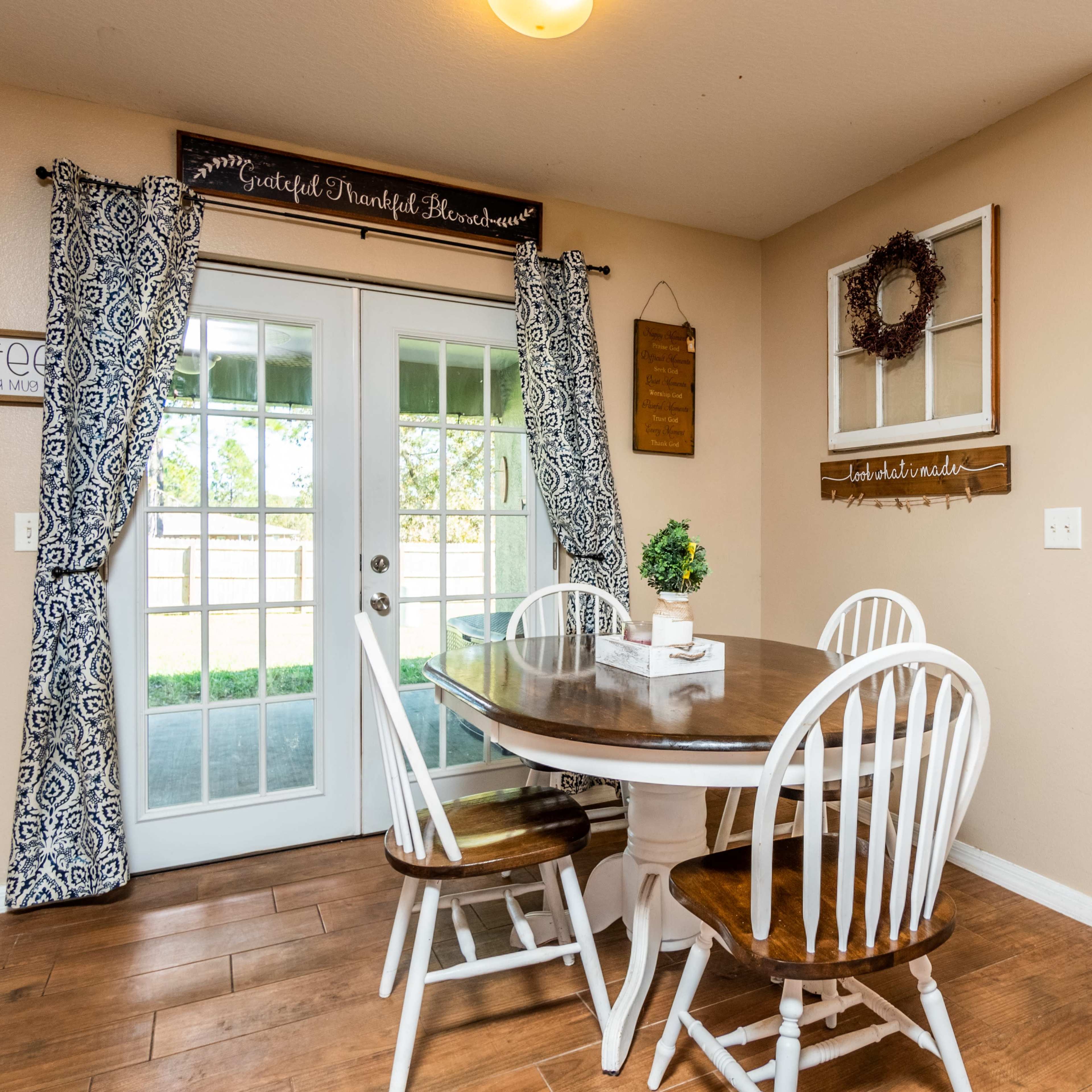 A dining area features a round wooden table with four chairs, surrounded by beige walls and large windows with patterned curtains.
