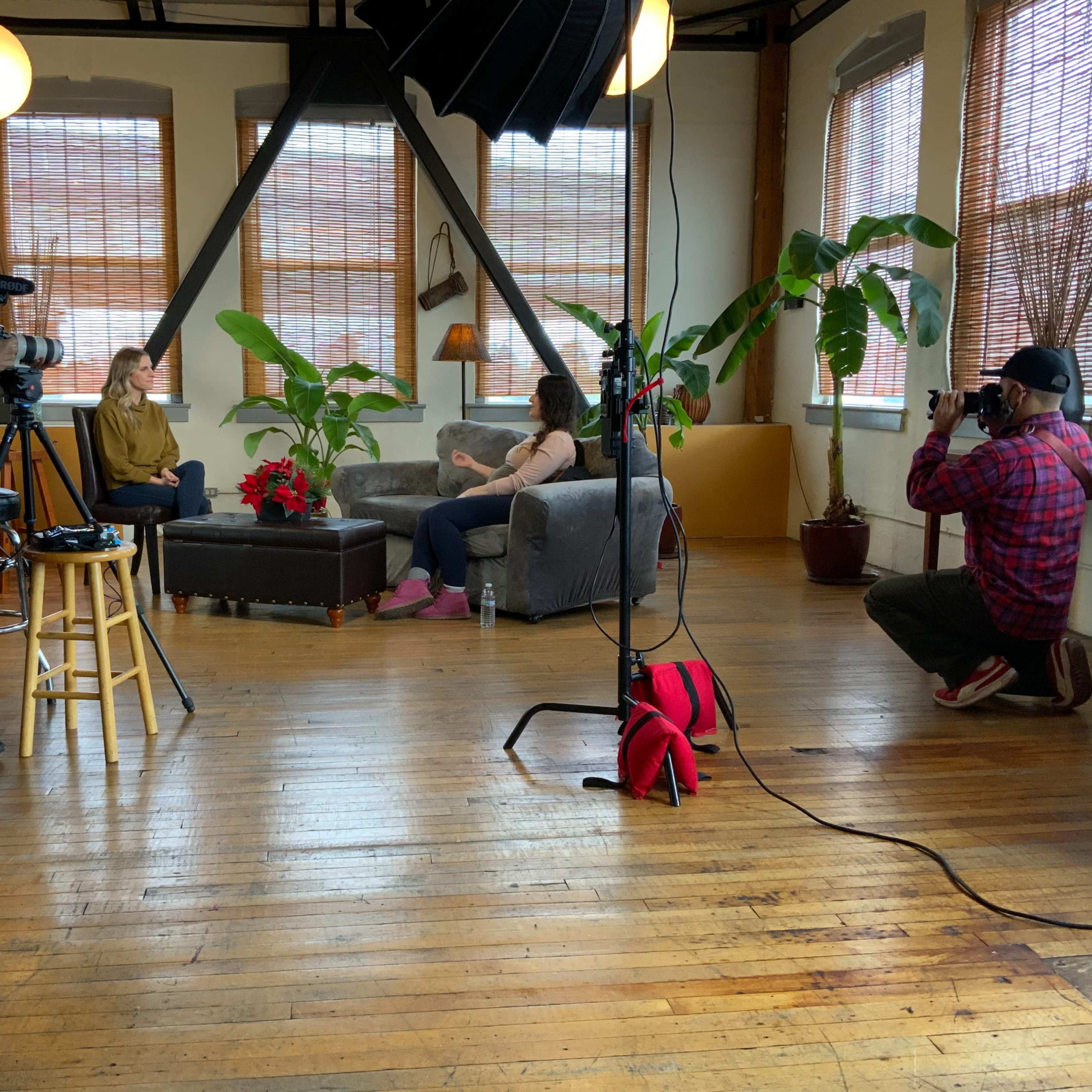 Two women sit on a couch in a well-lit studio while a camera crew captures the scene.