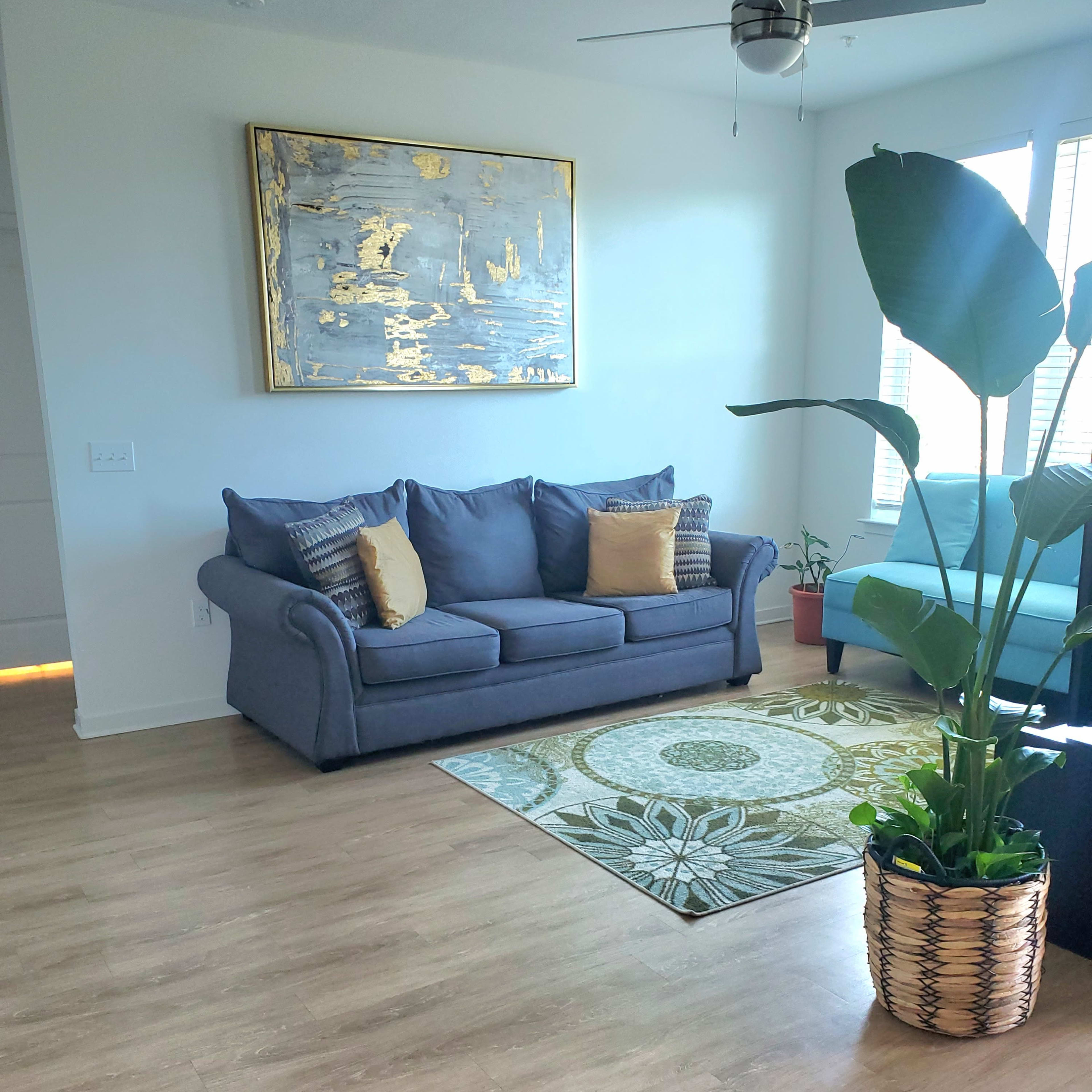 A living room with a blue sofa, a green armchair, a patterned area rug, a large decorative wall art piece, and a potted plant.